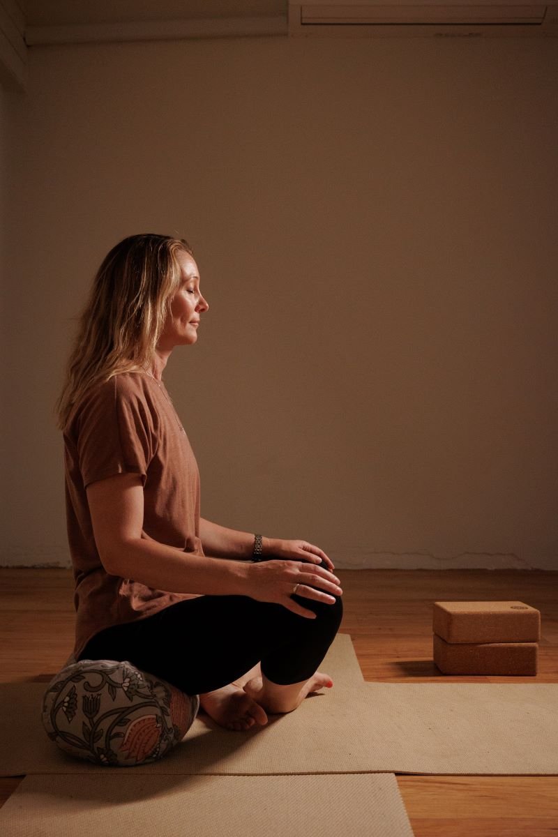 Woman practicing yoga or meditation in a serene indoor setting, sitting cross-legged on a yoga mat with a bolster under her knees, eyes closed, and hands resting on her knees.