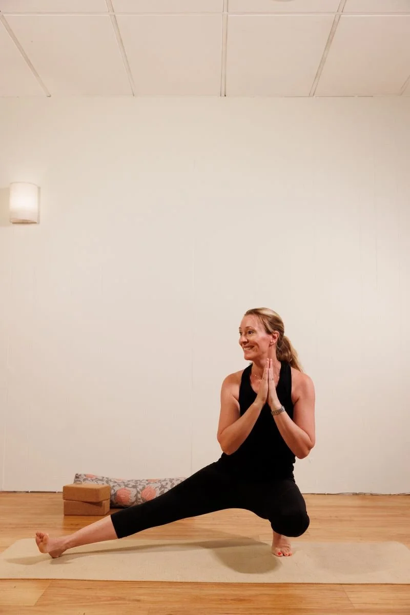 A woman practicing yoga indoors, kneeling on one leg with the other extended to the side, hands pressed together in a prayer position, smiling.
