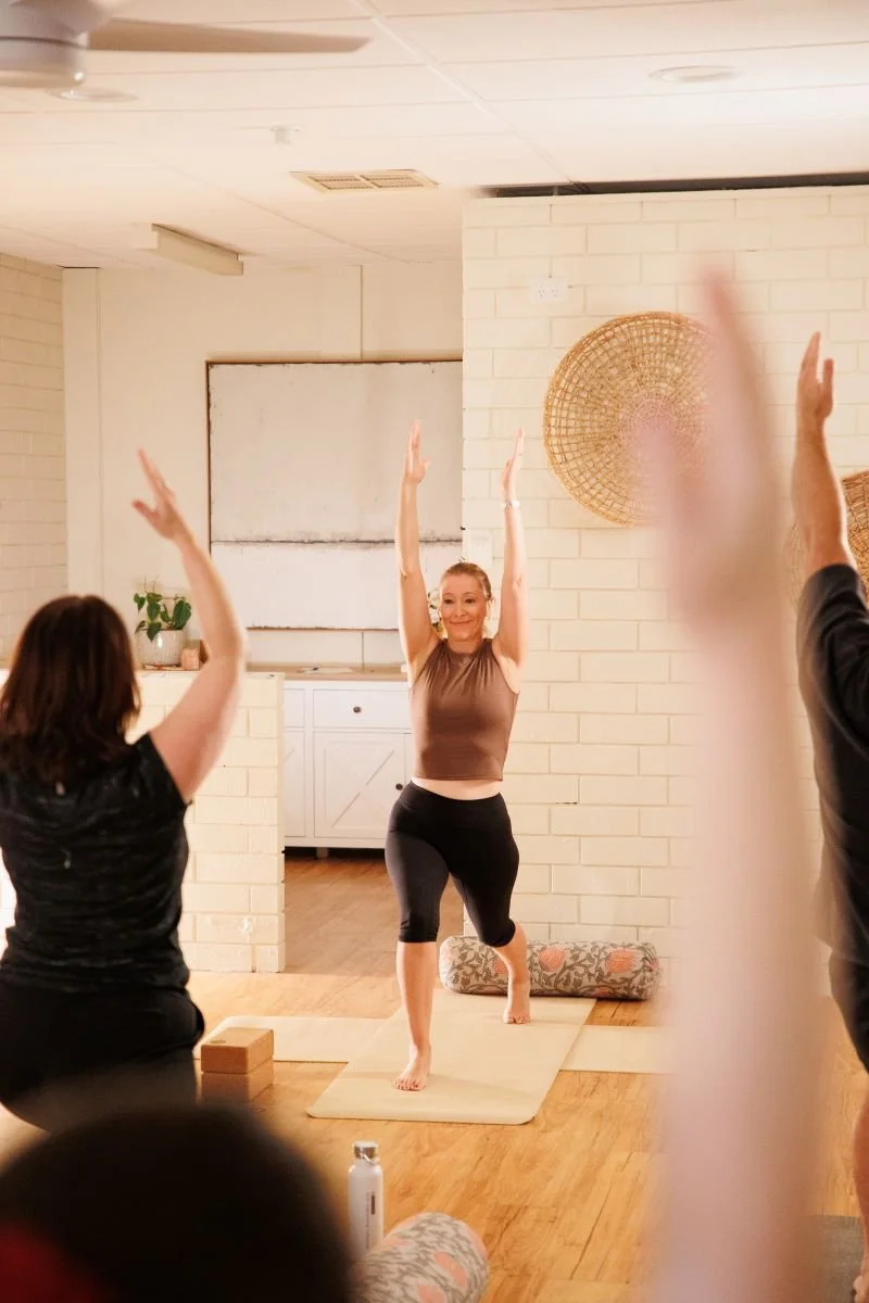 Women participating in a yoga or fitness class, guided by an instructor, in a cozy indoor setting with brick walls and decorative baskets.