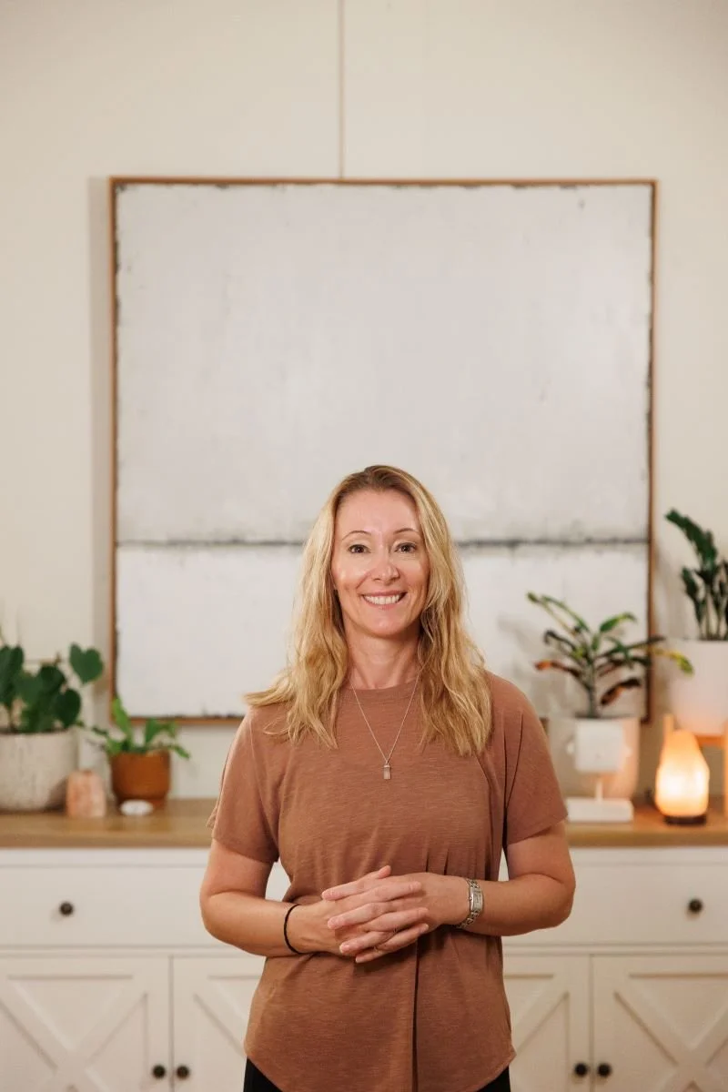 A woman with blonde hair smiling and standing in a room with a white cabinet, potted plants, and a large abstract painting in the background.