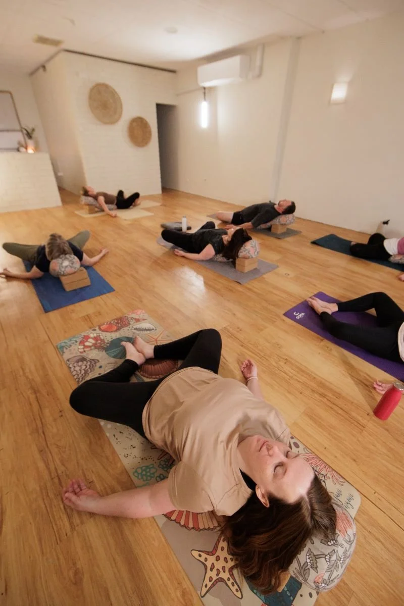 A group of people lying on yoga mats in a yoga or meditation class, with some using blocks for support, in a room with wooden flooring, wall decorations, and soft lighting.