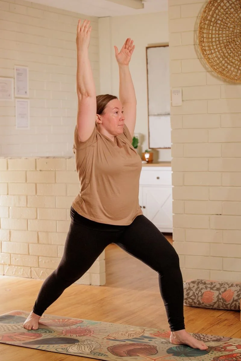 Woman practicing yoga in a spacious, well-lit room, standing in Warrior Pose on a colorful yoga mat with a brick wall background.