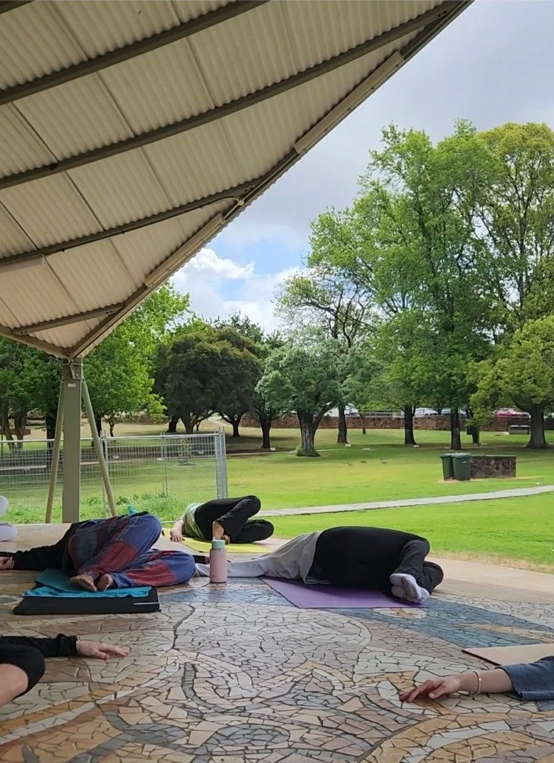 People practicing yoga on mats outdoors under a large shelter with a grassy park and trees in the background.