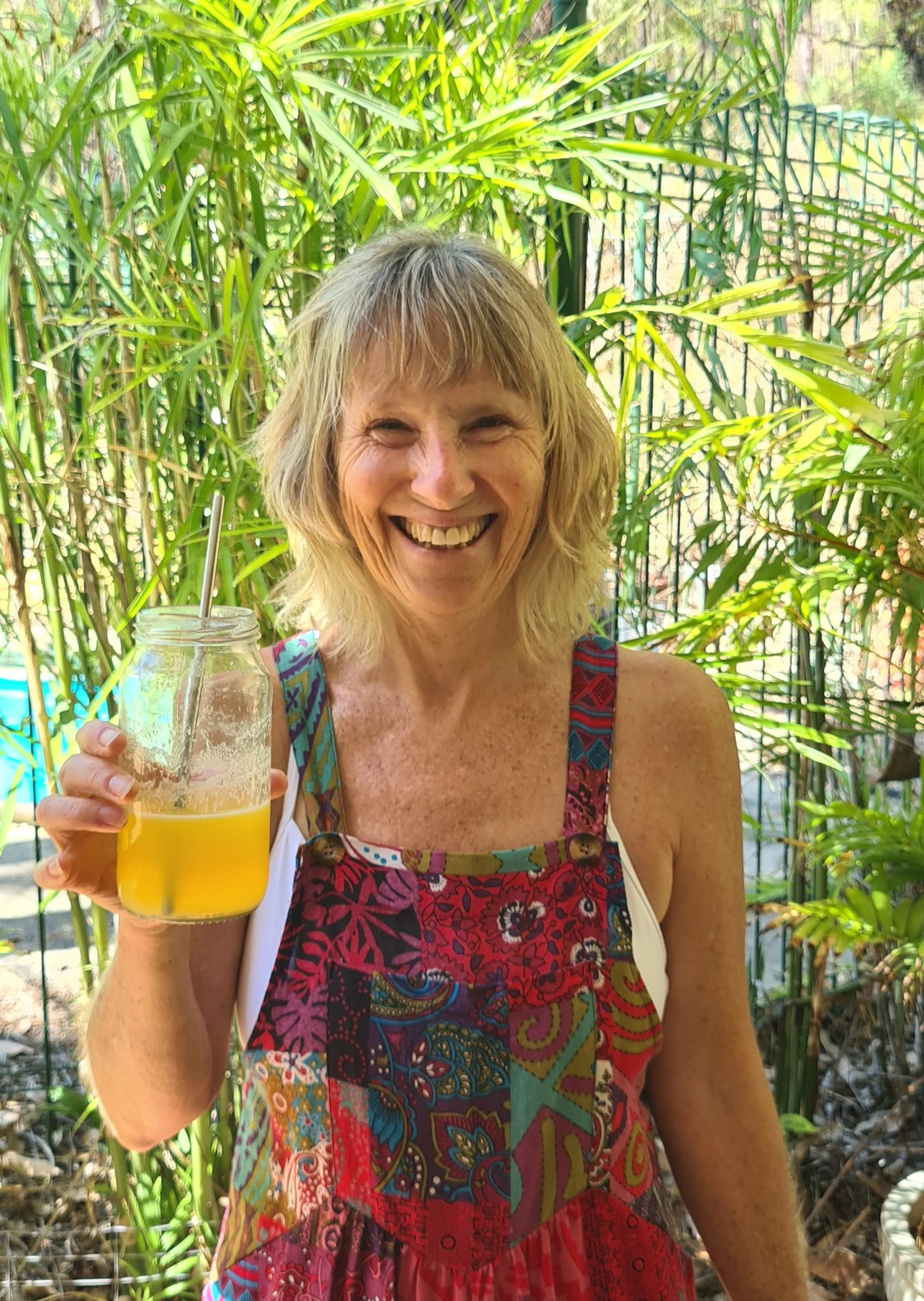 A smiling woman holding a glass of yellow beverage with a straw, outdoors amid green tropical plants.