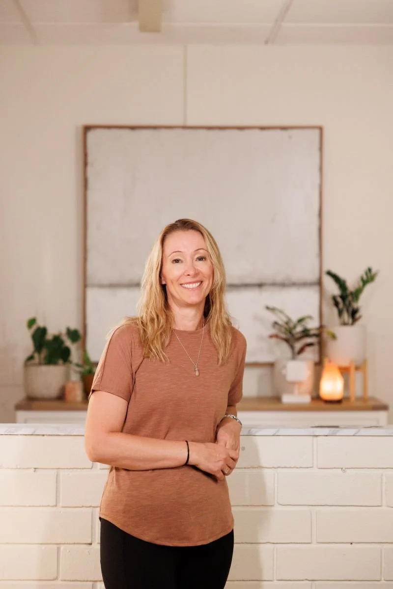 A woman with long blonde hair smiling while standing indoors, in front of a white brick wall with plants and decorations in the background.
