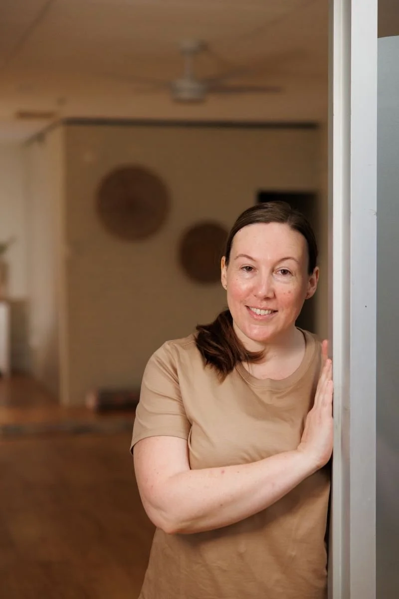 A woman with brown hair and freckles wearing a tan T-shirt, smiling, standing beside a door or wall, with a blurred background featuring decorative wall hangings.