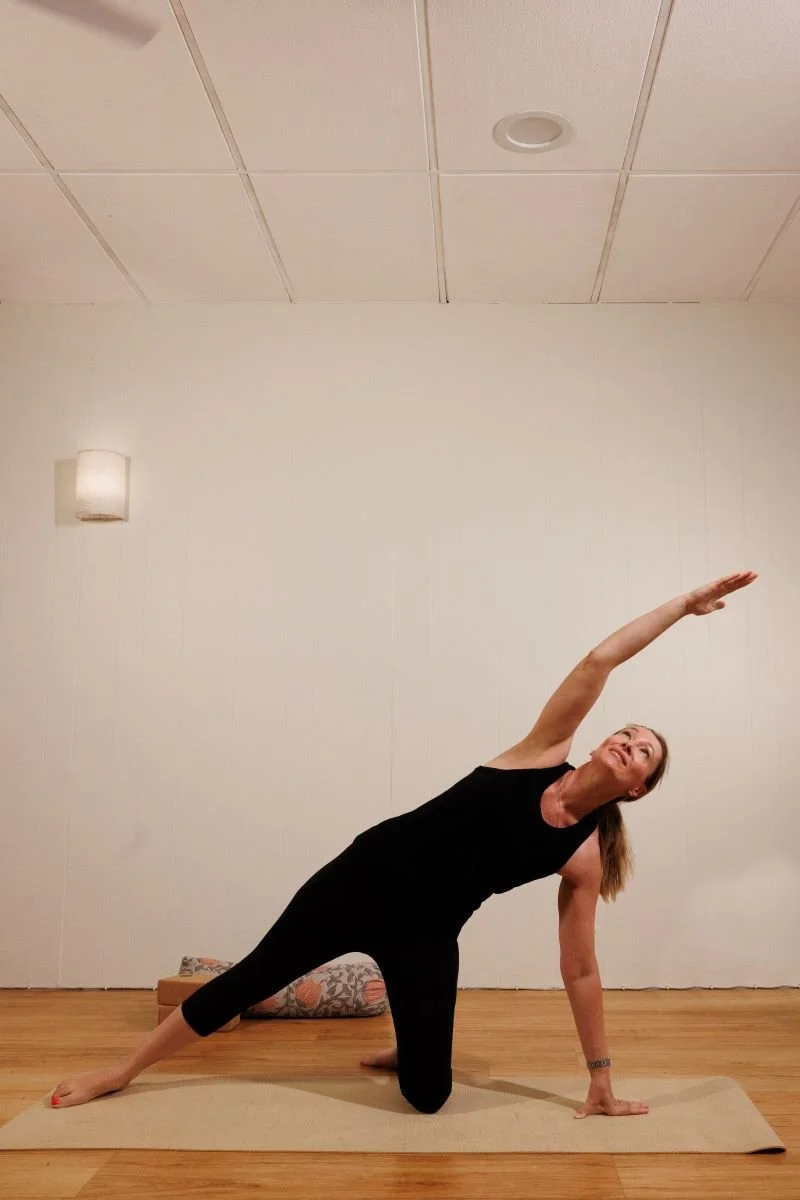A woman practicing yoga in a room with wooden flooring, doing a side plank pose with one arm extended upward, wearing a black sleeveless top and black leggings.
