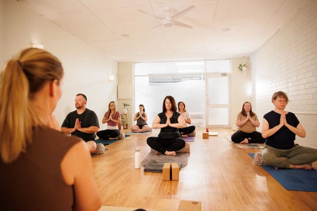People participating in a yoga or meditation class, sitting cross-legged on mats with hands in prayer position, in a bright room with wooden floors and white brick walls.