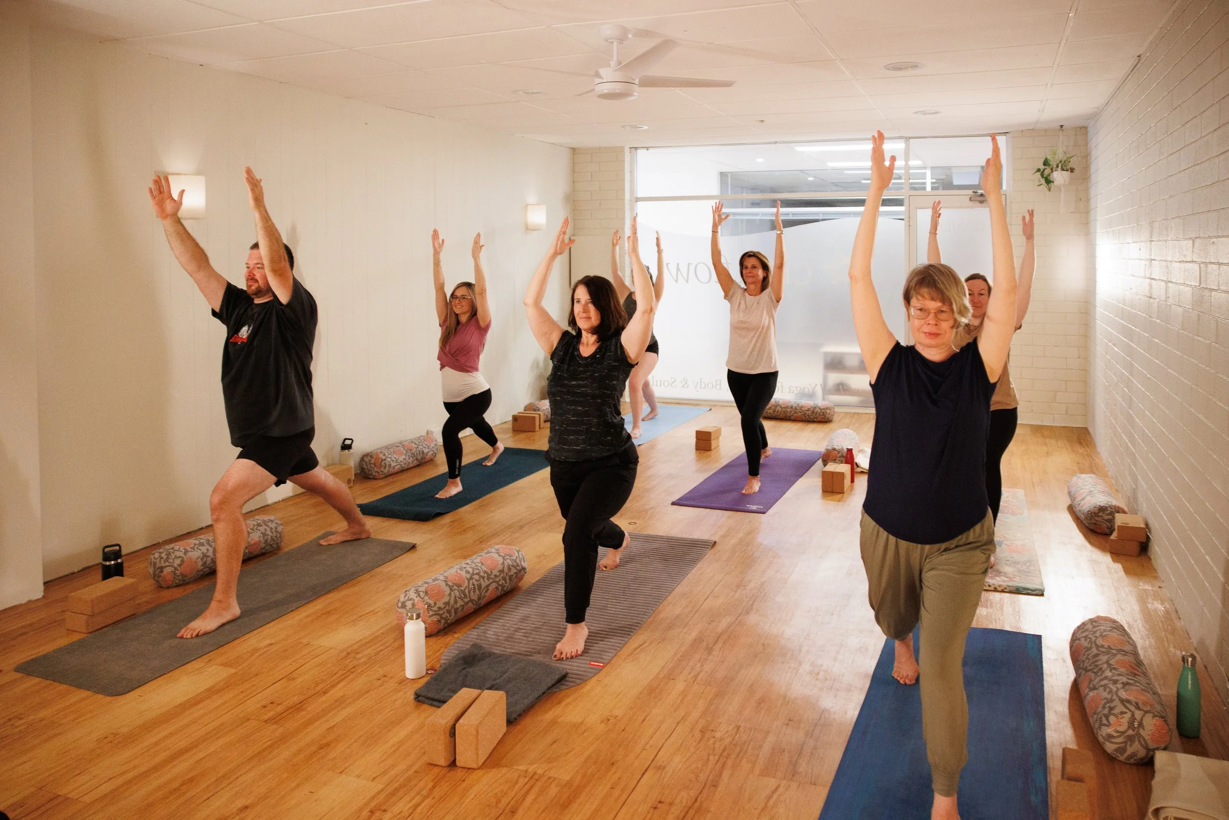 A group of people participating in a yoga class in a bright, indoor studio, standing on yoga mats with their arms raised above their heads.