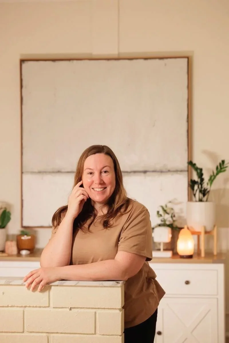 Woman with long brown hair smiling, leaning on a white brick surface in a cozy room with plants, lamps, and a large abstract artwork in the background.