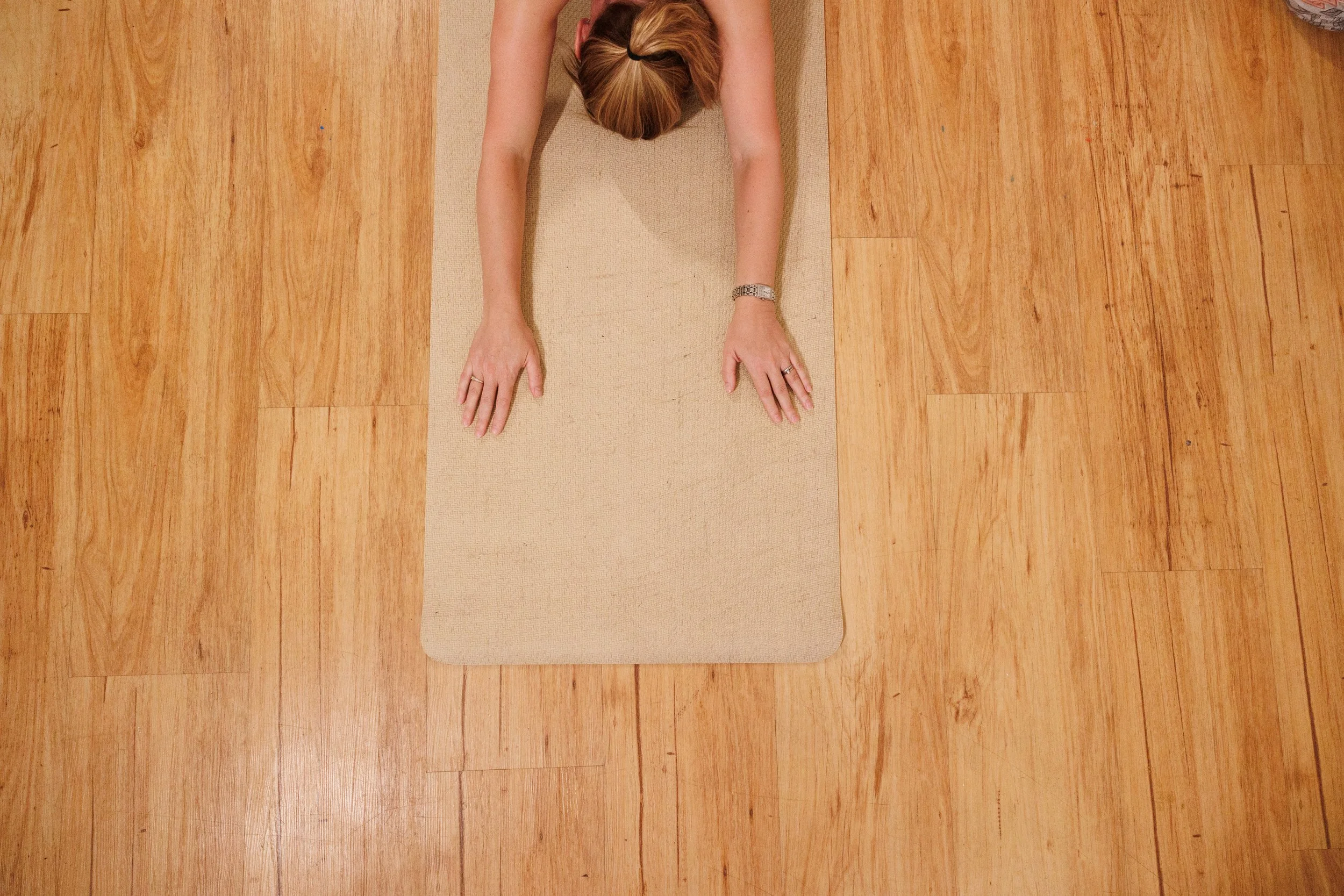 Top-down view of a woman performing yoga on a beige mat on a wooden floor, her arms extended forward and head resting on the mat, in a stretching pose.