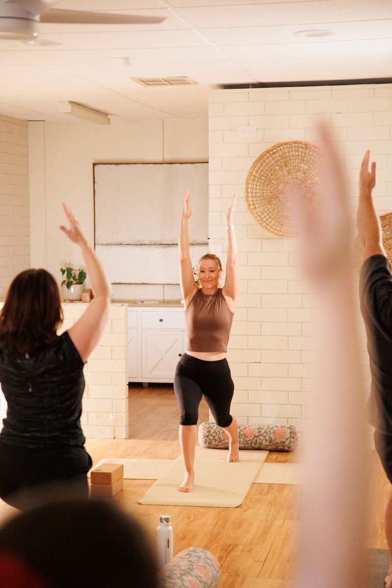 Women practicing yoga in a studio, with one woman standing on a yoga mat and others raising their arms.