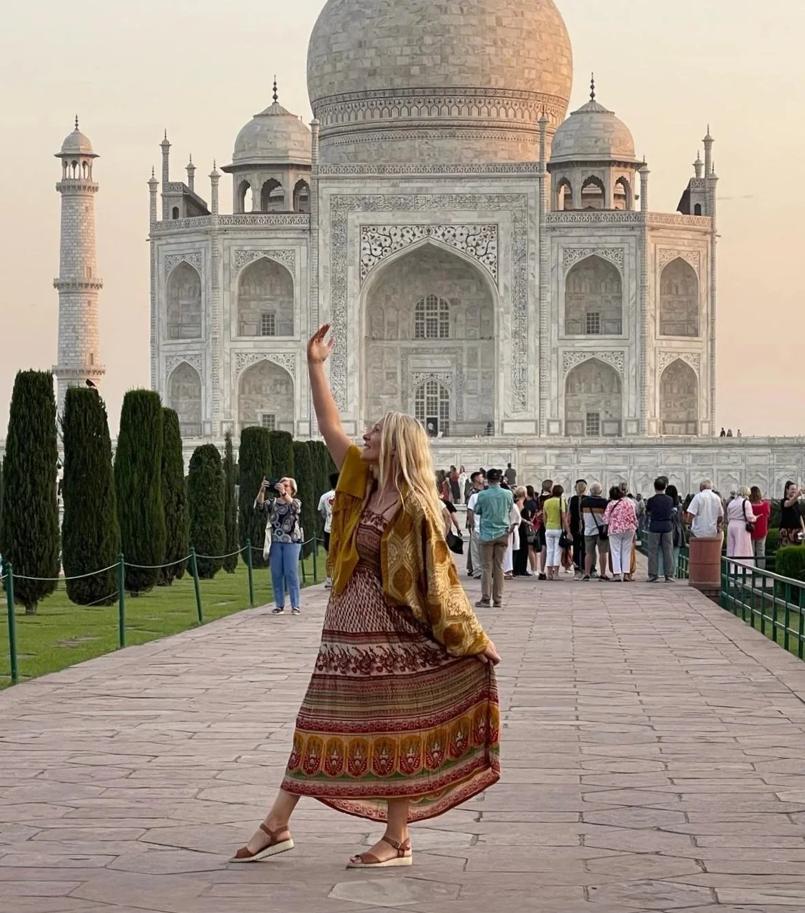 A woman in a patterned dress and yellow jacket posing in front of the Taj Mahal, with other tourists in the background and the monument's white marble structure visible at sunset.