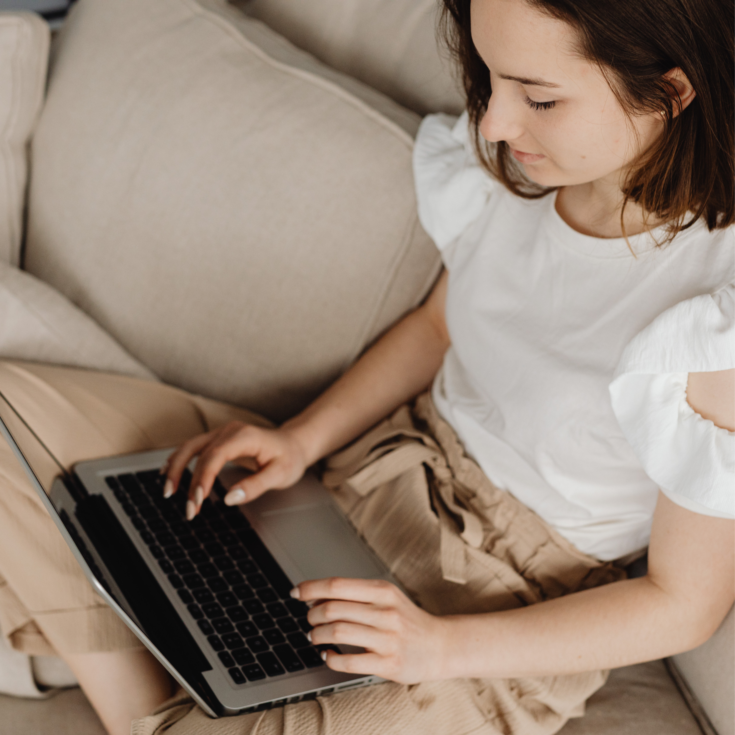 A young woman with brown hair wearing a white shirt sitting on a beige couch, using a laptop with a black keyboard.