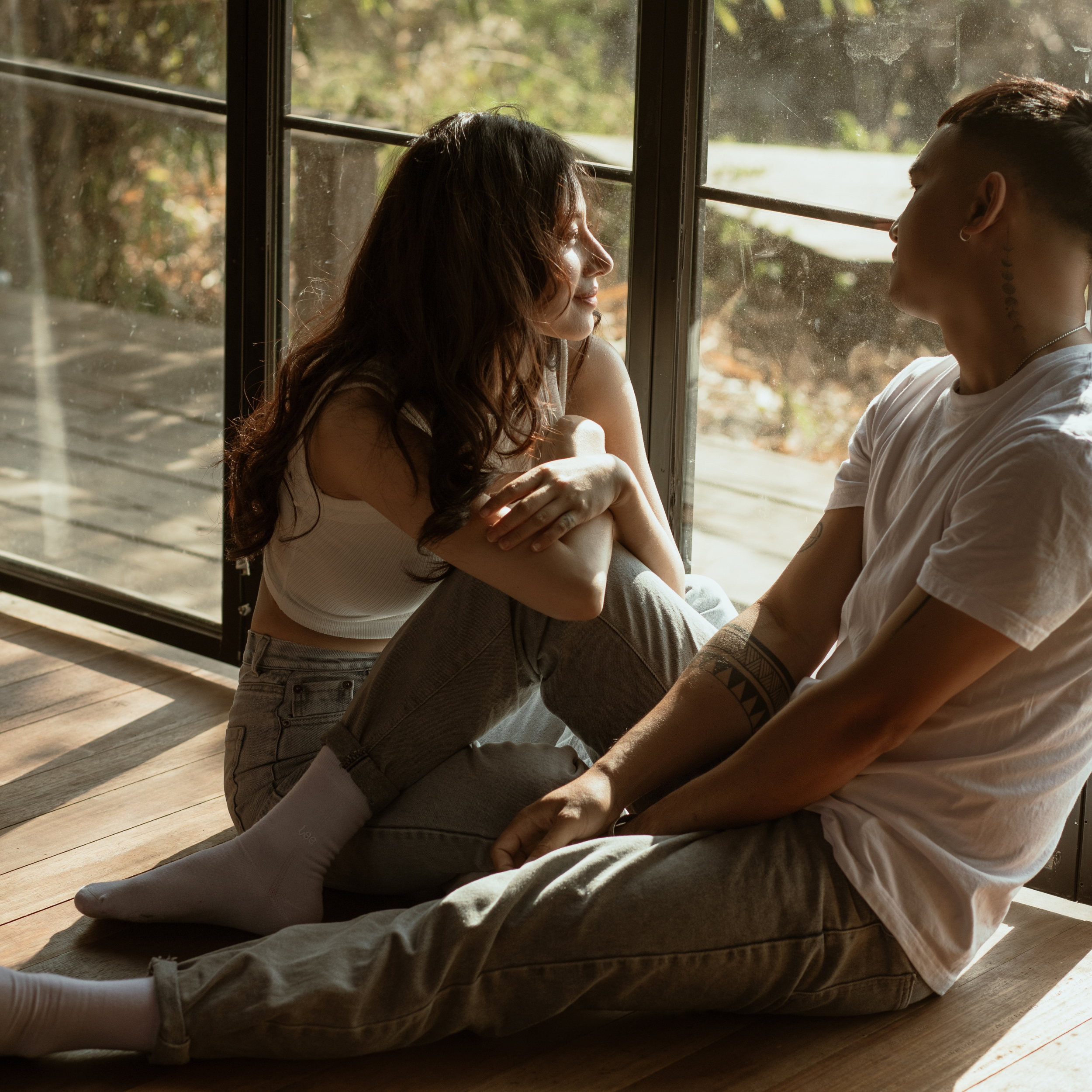 Two people sitting face to face on the floor near a large window with sunlight streaming in, engaging in a close and intimate conversation.