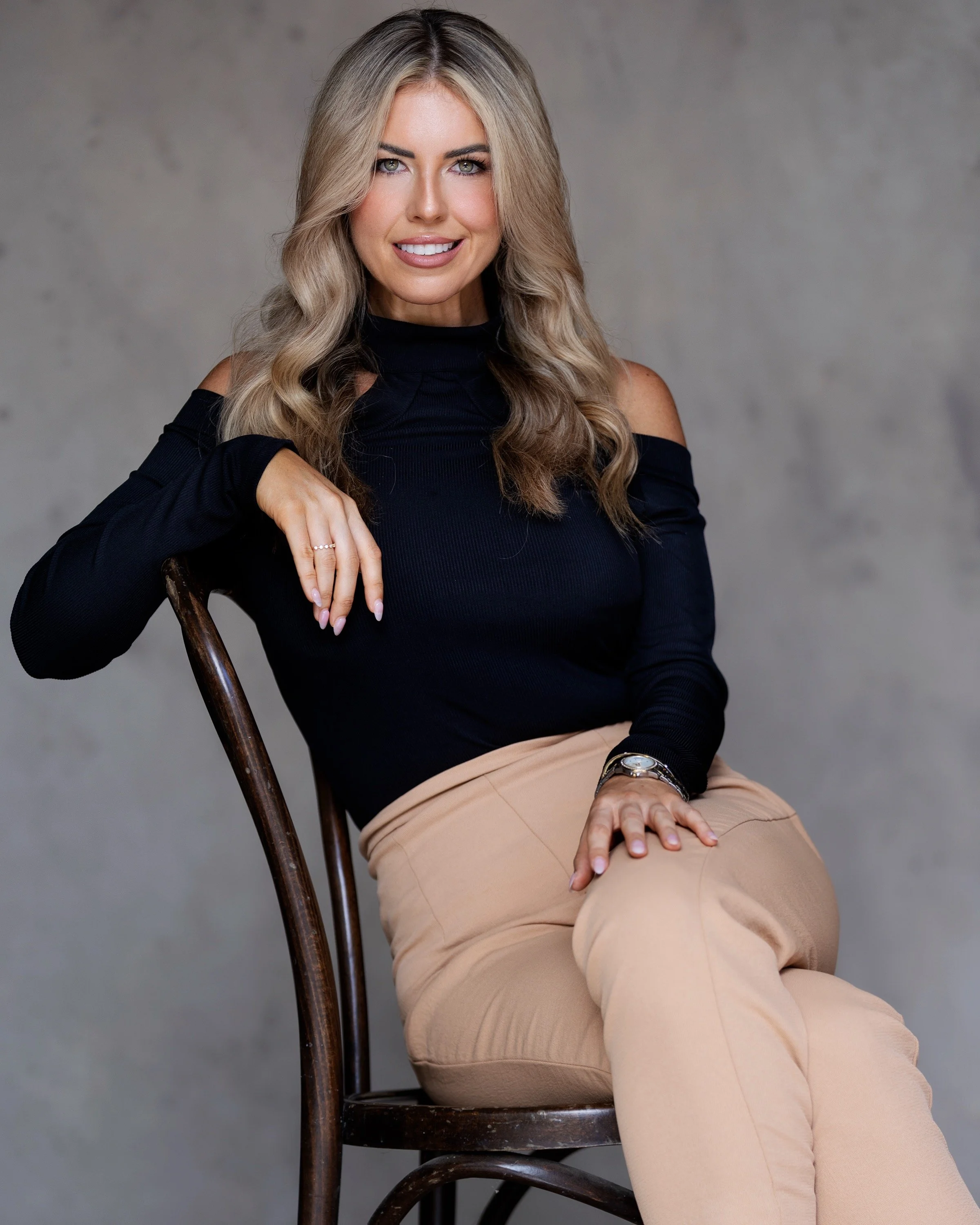 A woman with wavy blonde hair wearing a black off-the-shoulder top and beige pants, sitting on a wooden chair against a plain gray background.