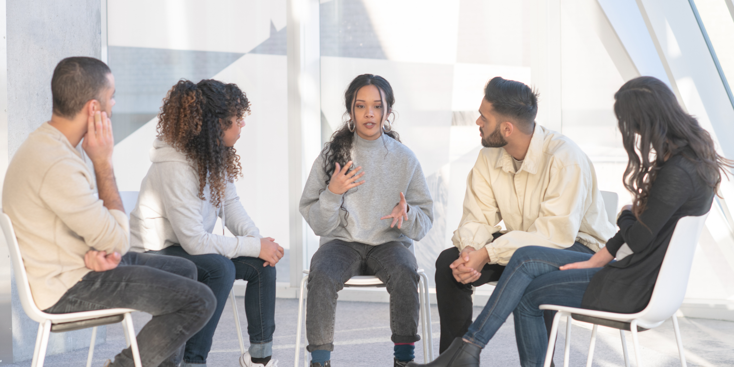 A woman sitting in the middle and speaking to a group of five diverse people seated in a circle inside a bright room.