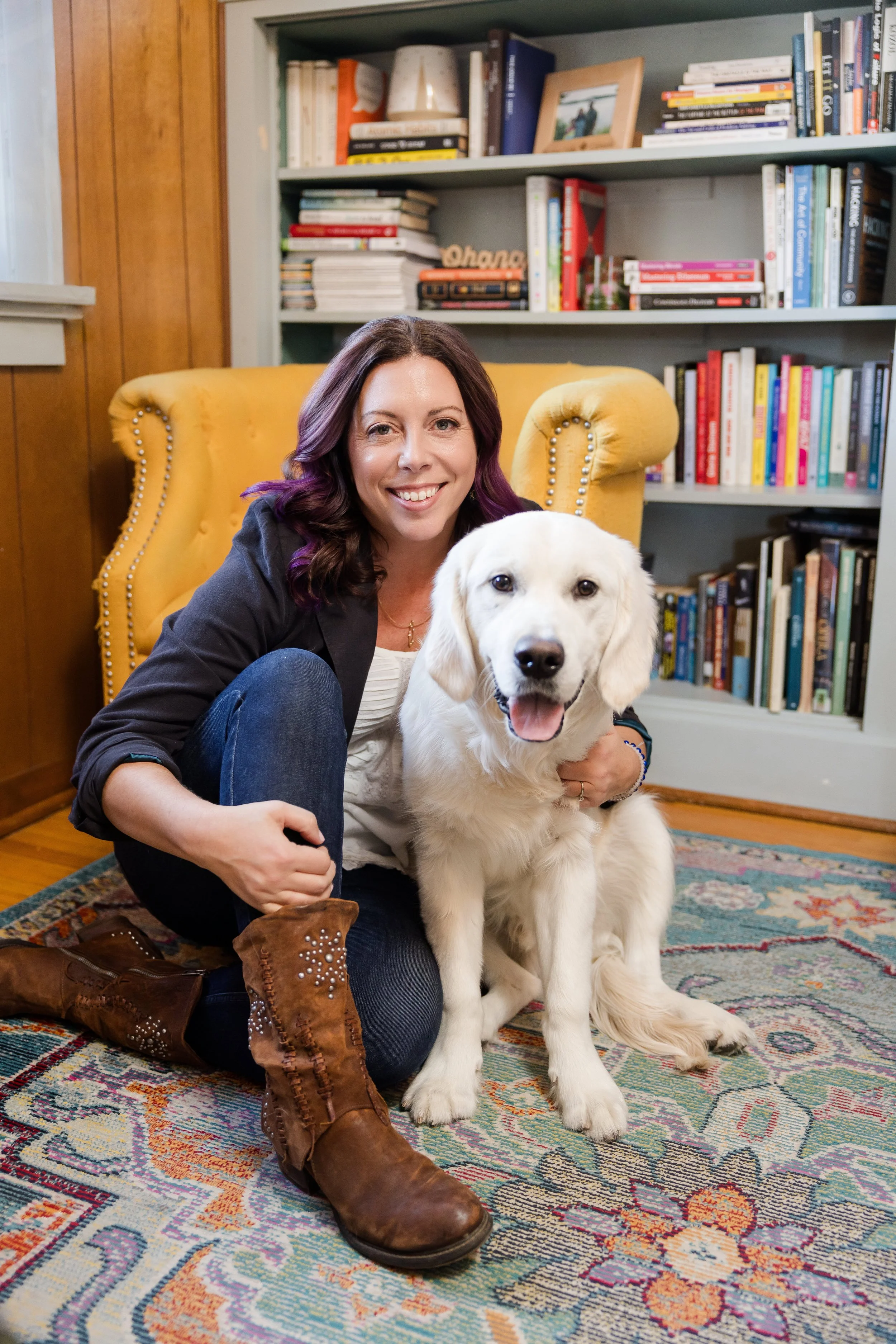 A woman sitting on a colorful rug with a yellow armchair behind her, holding a happy Labrador retriever puppy inside a home library with shelves of books.