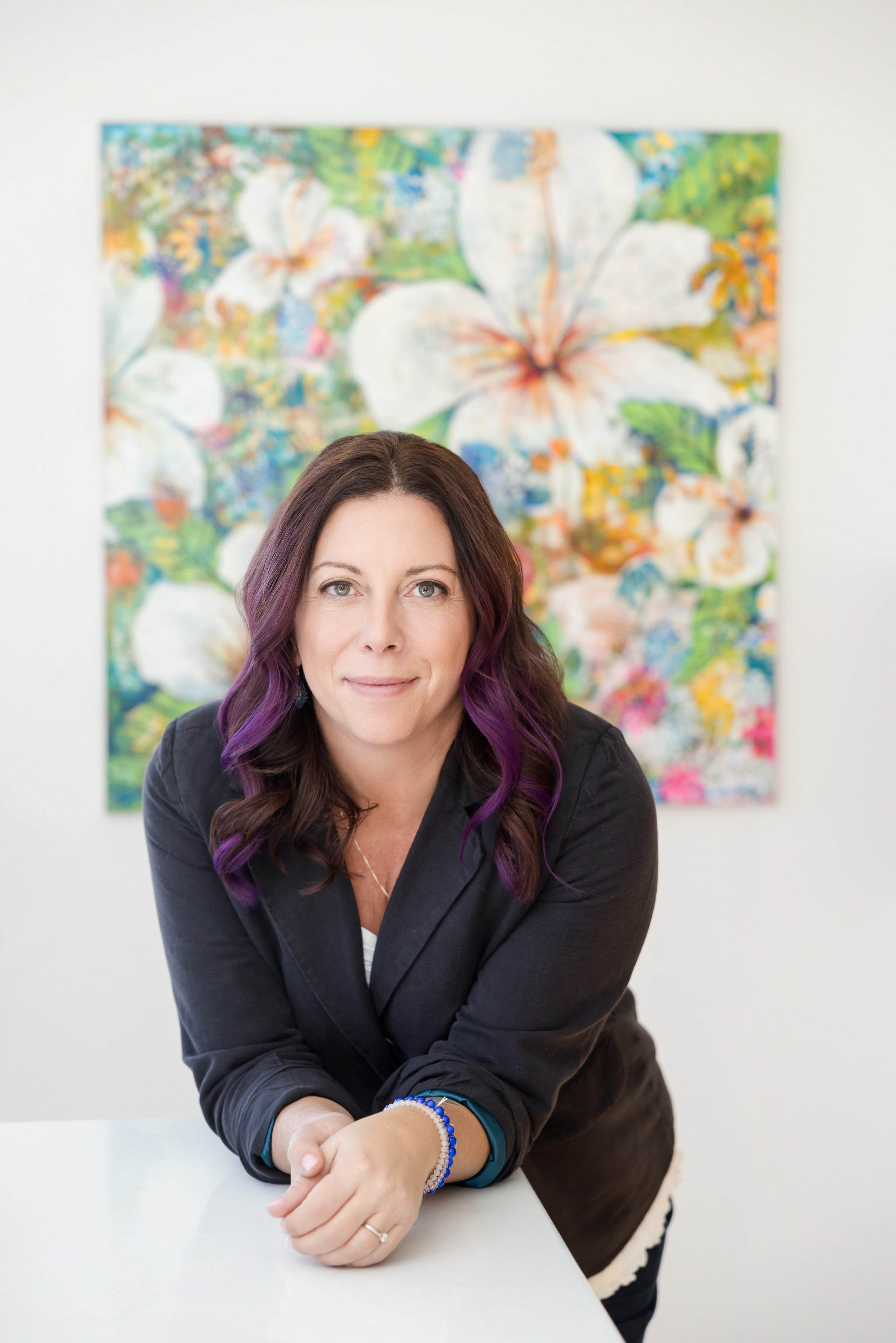 A woman with purple and brown hair, smiling and leaning on a white table in front of a colorful floral painting.