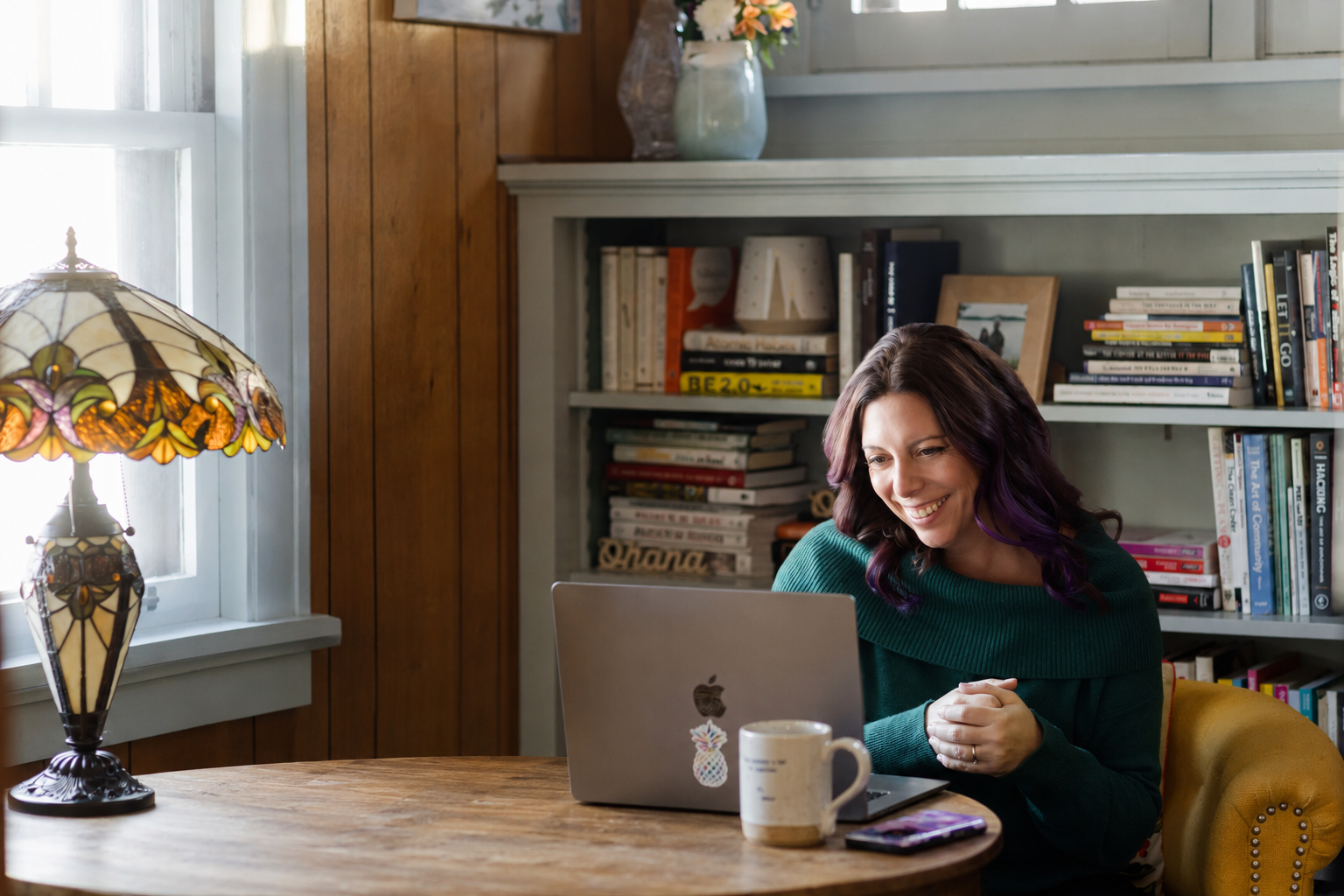 A woman with purple hair sitting at a wooden table in a cozy room, using a silver MacBook laptop. The room has wooden and white walls, a bookshelf filled with books, a colorful stained glass lamp, a mug, and a smartphone on the table.