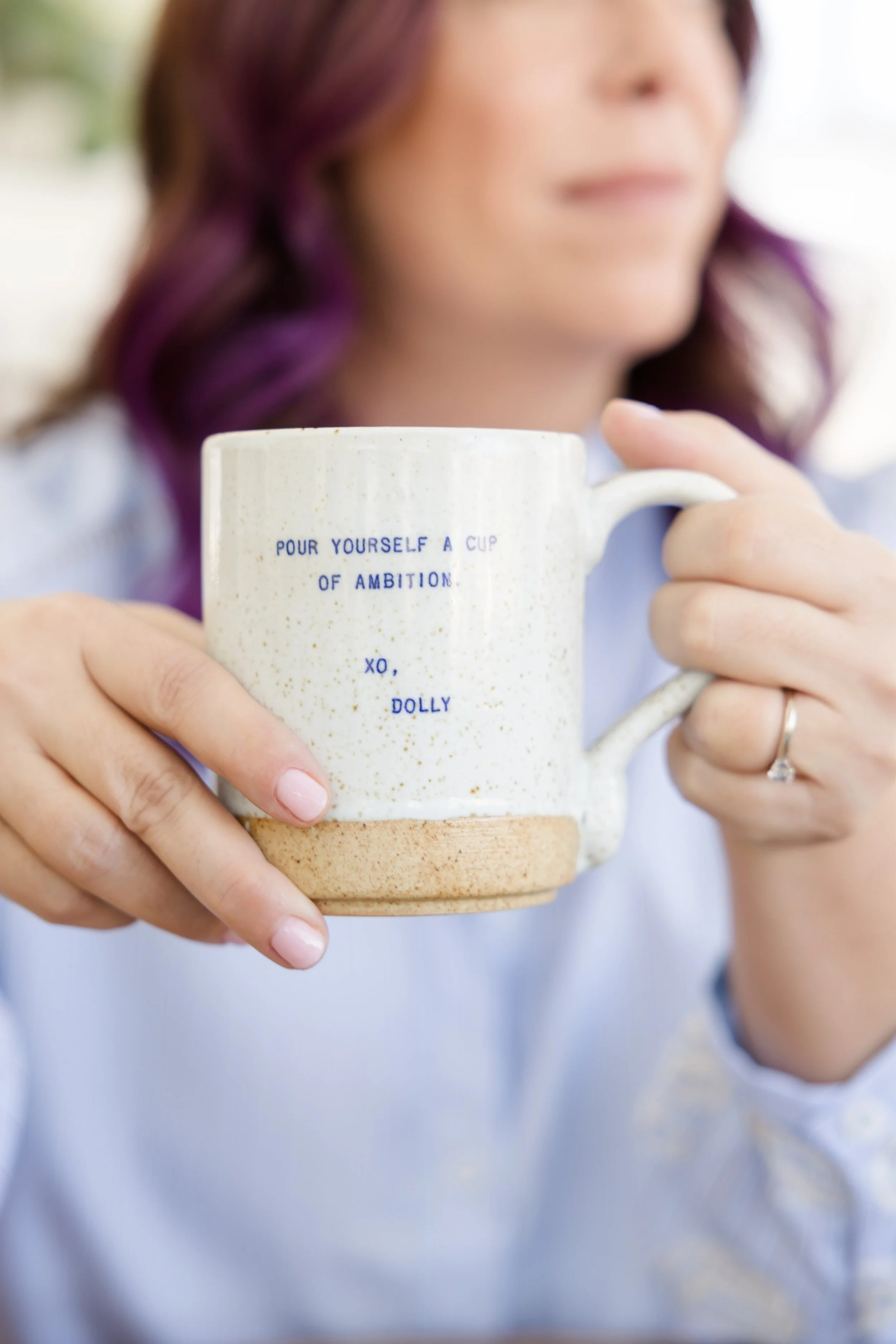 A woman with purple hair holds a beige and white speckled ceramic mug with a message that says, 'Pour yourself a cup of ambition. Xo, Dolly'.
