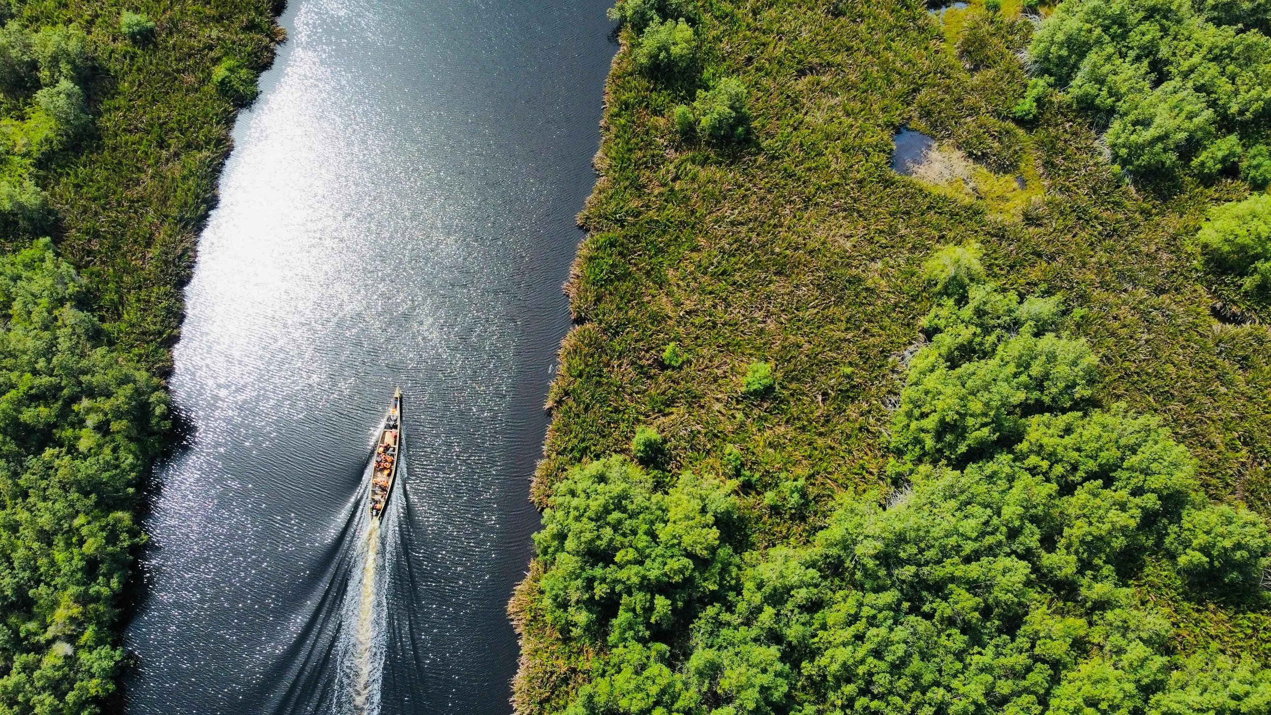 Aerial view of a boat traveling through a narrow river or canal surrounded by dense green forest.