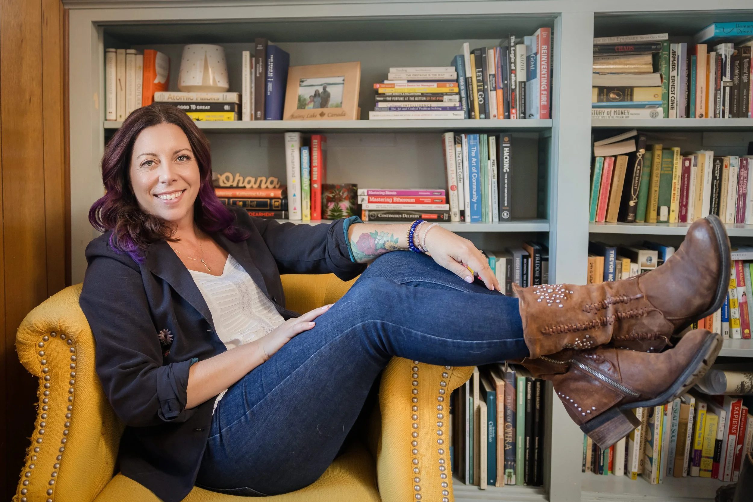 A woman with brown hair and purple tips, sitting on a yellow armchair in front of a bookshelf, smiling at the camera, wearing a black jacket, white top, blue jeans, and brown boots, with her right arm resting on the armrest and her left leg crossed over her right.