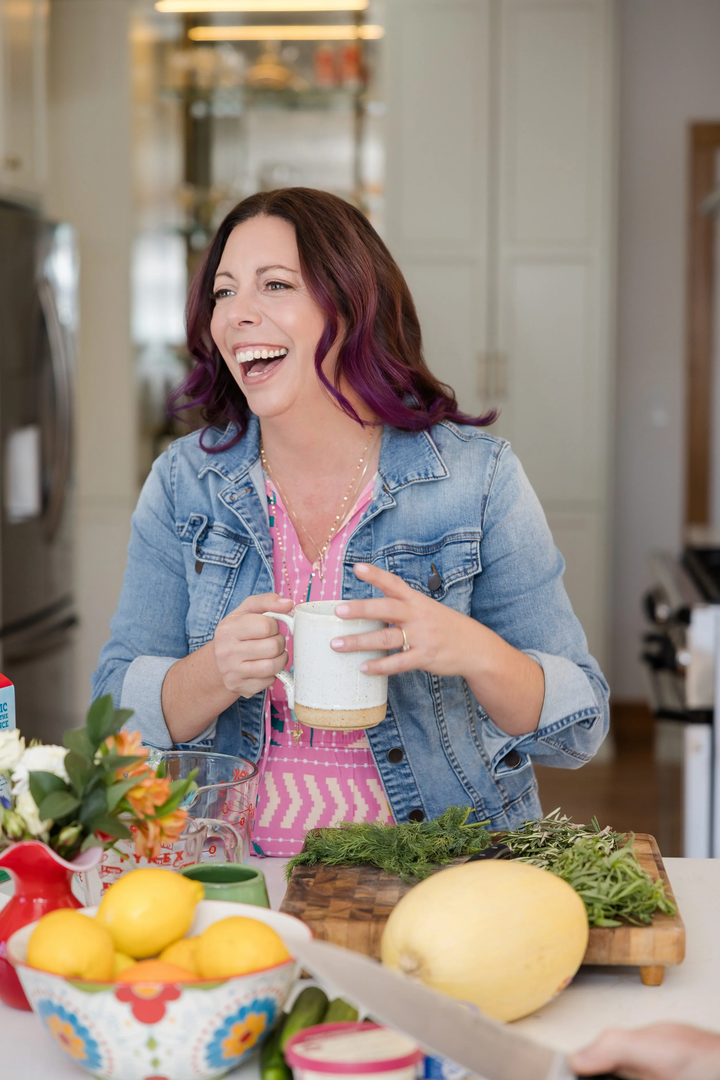 A woman with purple hair and a denim jacket is smiling and holding a mug in a kitchen. The counter in front has various fruits and vegetables, including lemons, a melon, and leafy greens.