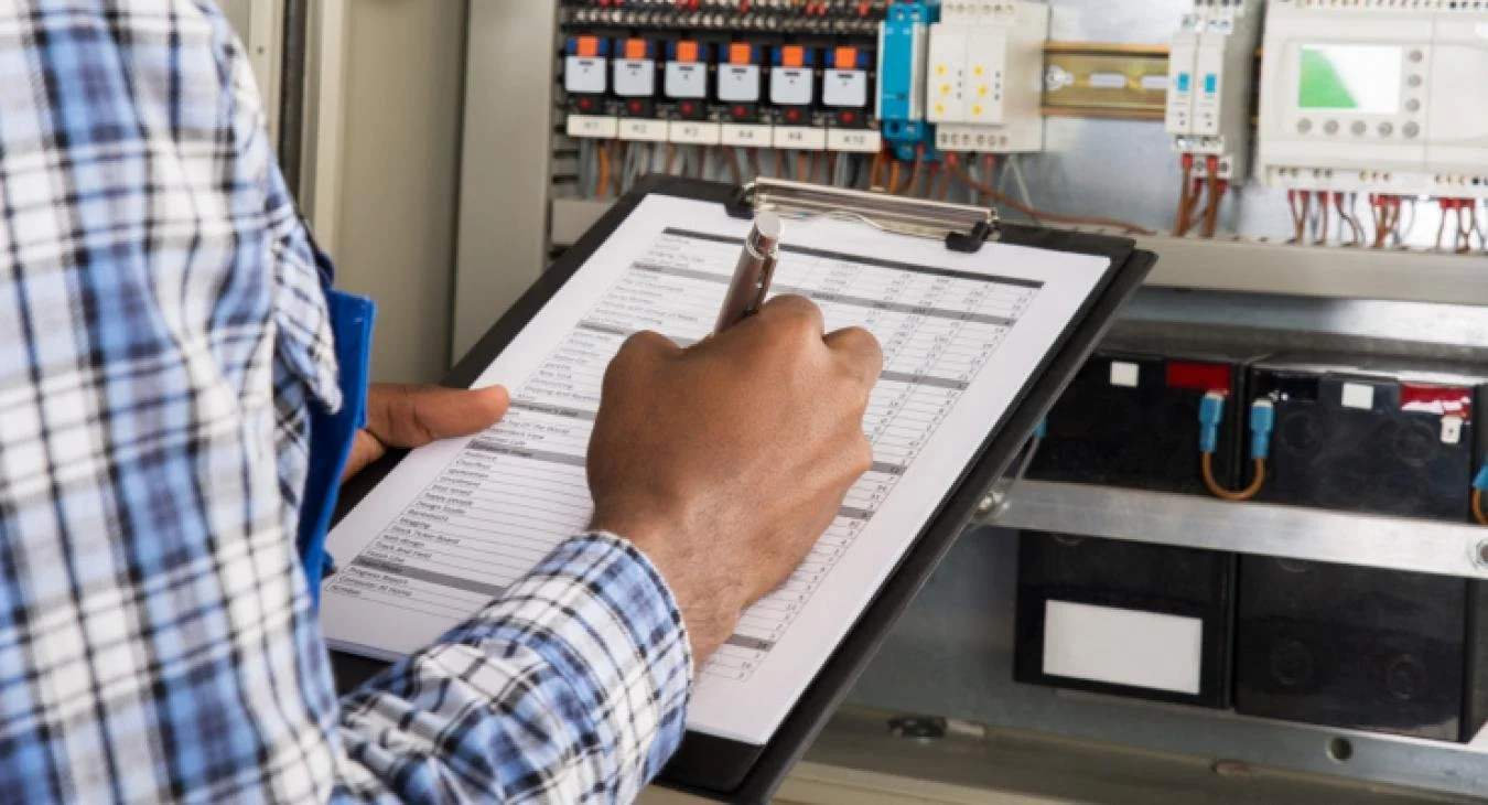 Person using a clipboard to check electrical equipment and wiring in an electrical control panel.