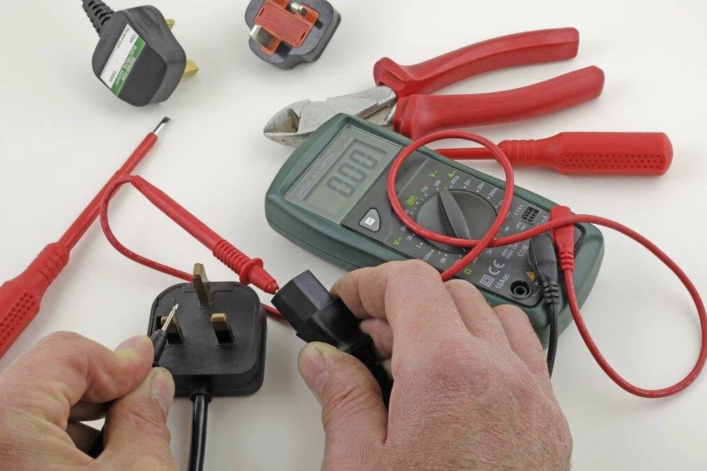 Person using a multimeter to test an electrical power outlet, with various electrical tools and adapters on a white surface.
