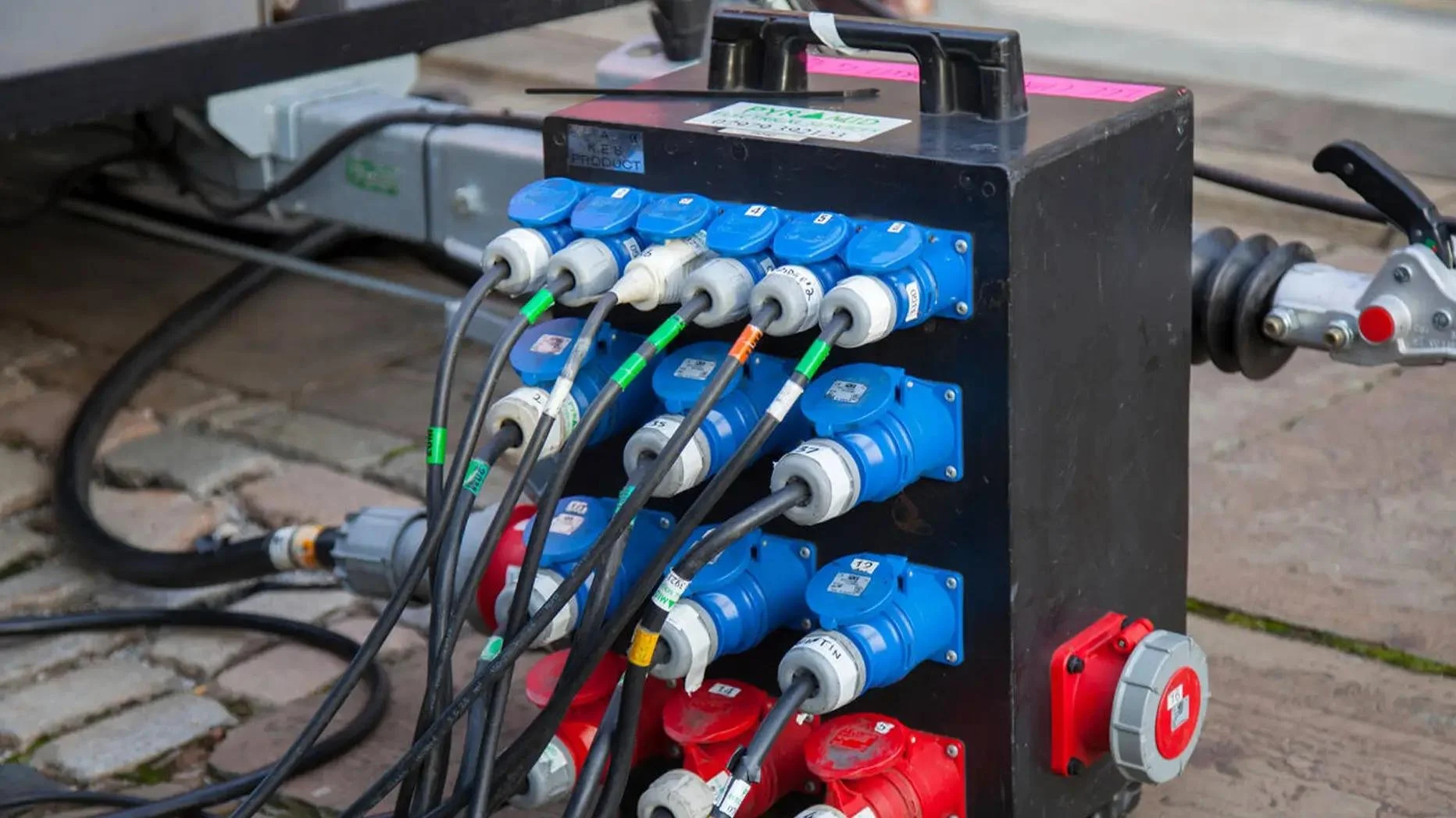 A black electrical box containing multiple blue, red, and gray electrical connectors and cables connected to it, placed on a brick surface outdoors.