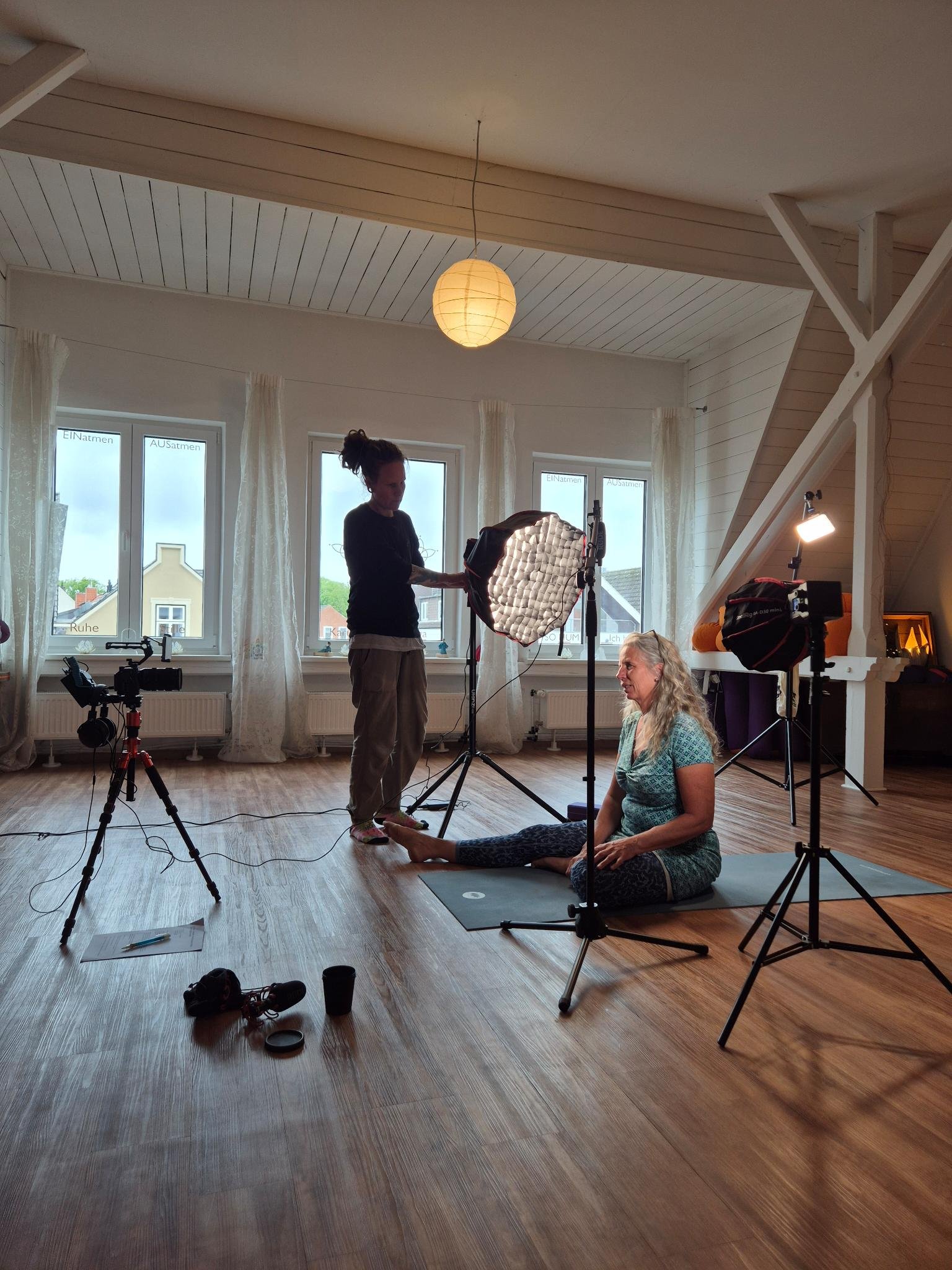 A woman sitting on the floor during a photoshoot or video filming session with professional lighting equipment. A person standing behind her adjusts the lighting, and various camera gear and accessories are scattered around on the wooden floor.