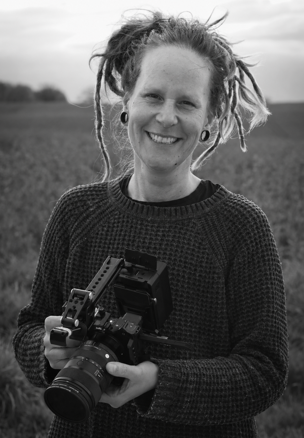 A woman with dreadlocks smiling outdoors while holding a professional camera with a stabilizer.