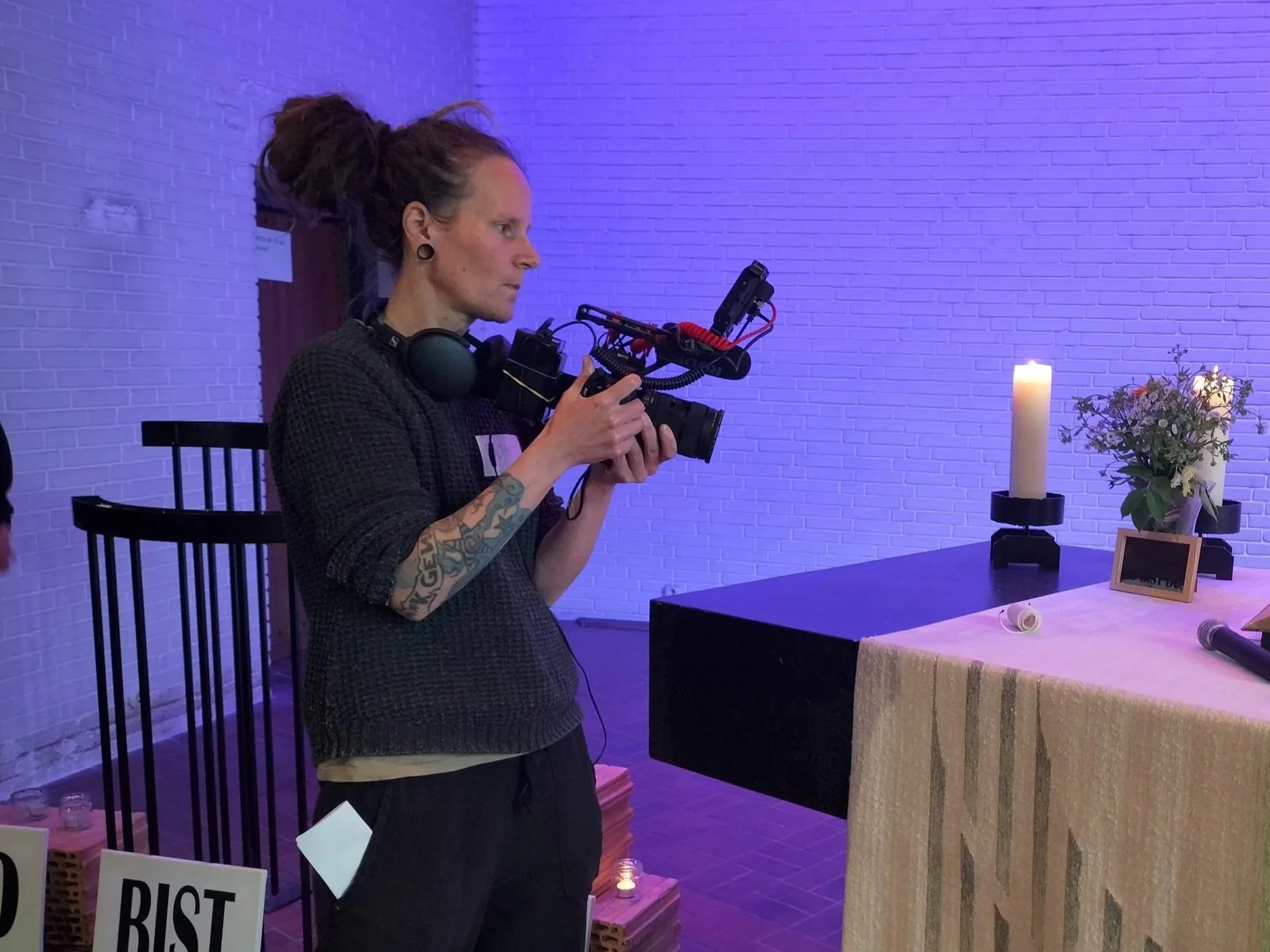A woman with tattoos on her right arm and dark hair in a bun operating a camera, standing next to a table with a lit candle, flowers in a vase, and framed photographs, in a room with purple lighting.
