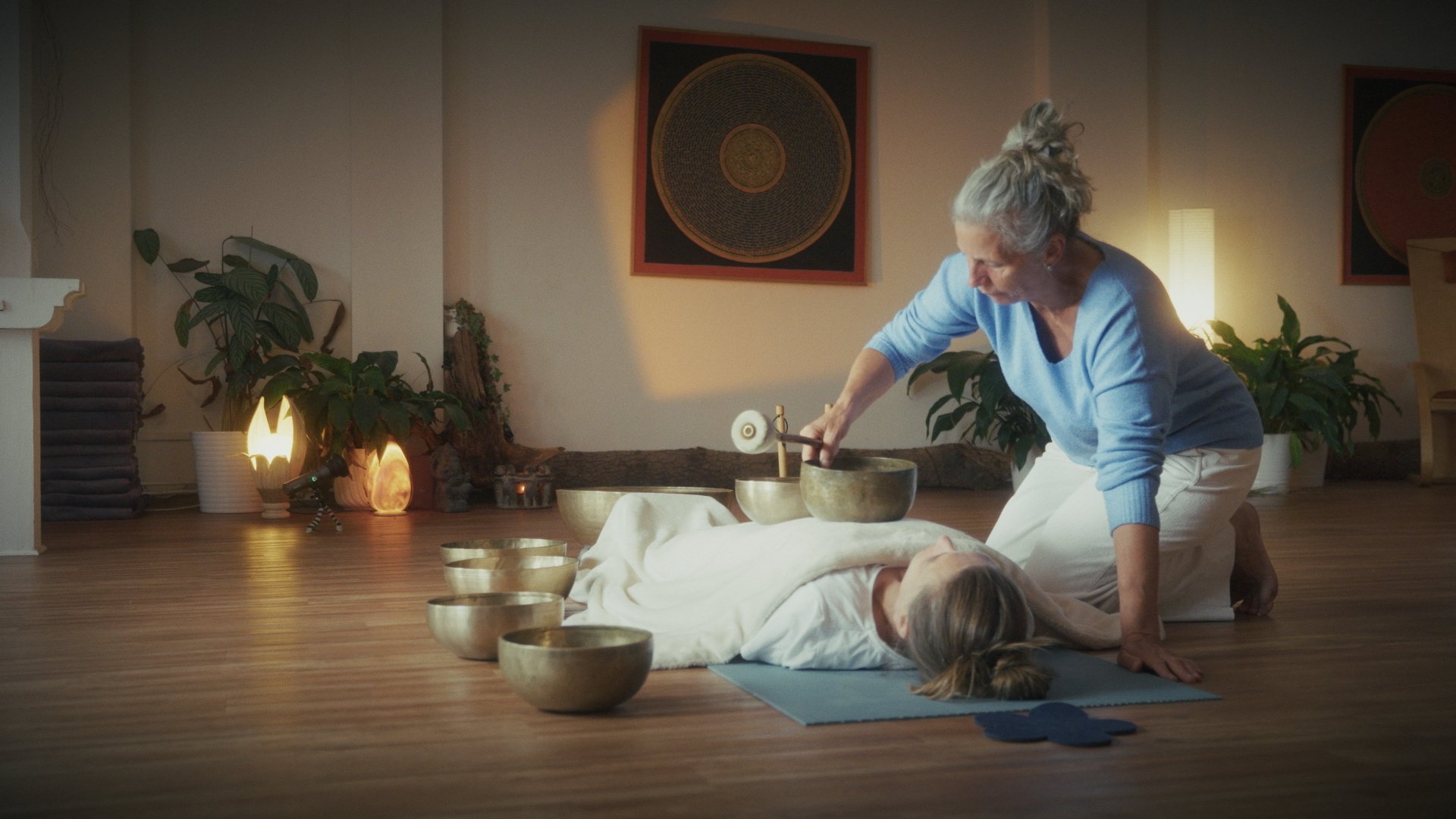 A woman practicing sound healing or meditation with singing bowls in a calm, warmly lit room filled with plants and artwork.
