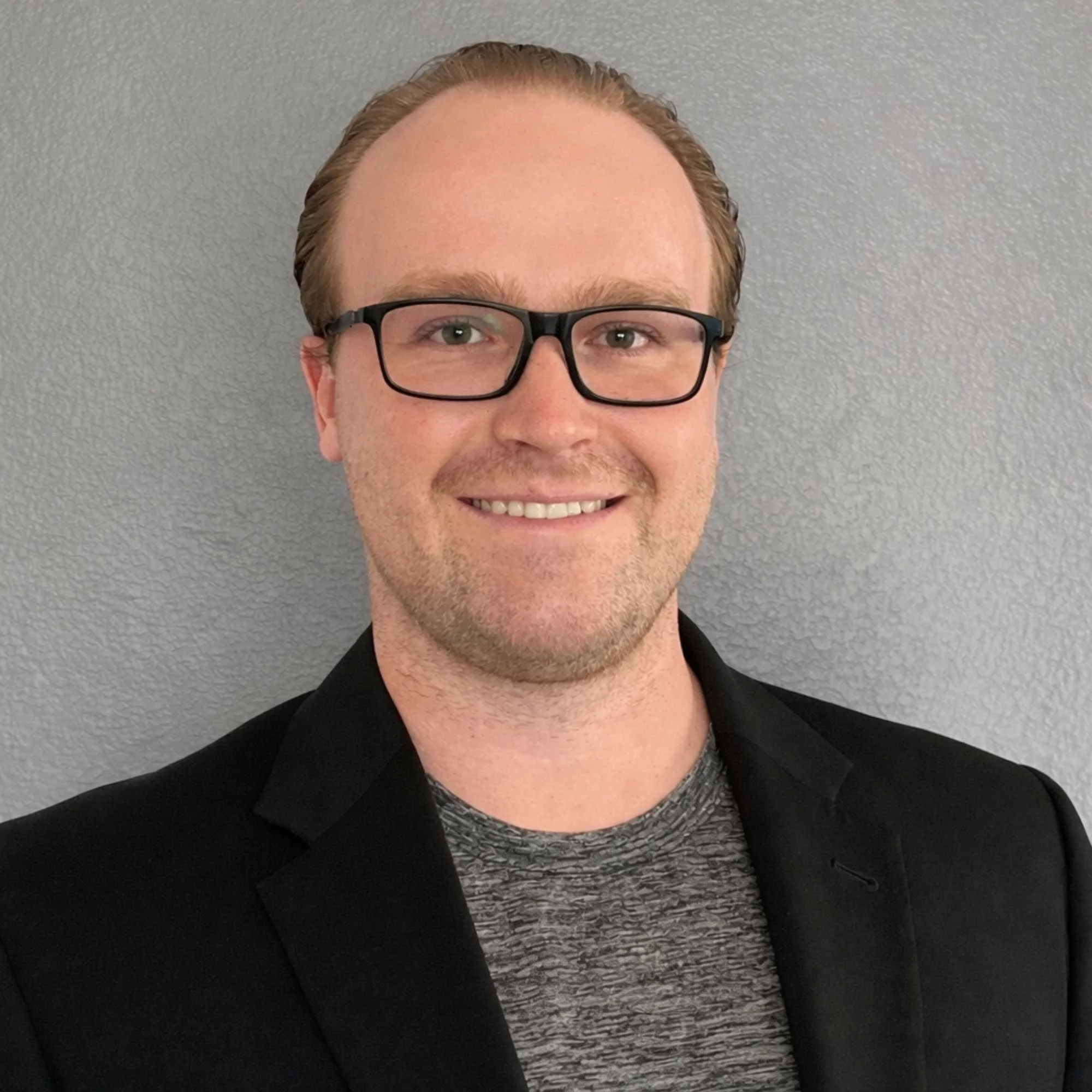 A portrait of Adam smiling, wearing glasses, a black blazer, and a patterned shirt, standing against a plain, light gray textured wall.