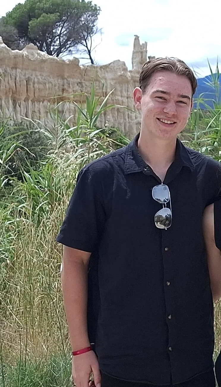 Jongen met bruin haar, zwart overhemd en zonnebril hangt aan zijn shirt, glimlachend op een natuurlijke achtergrond met gras en rotsen.