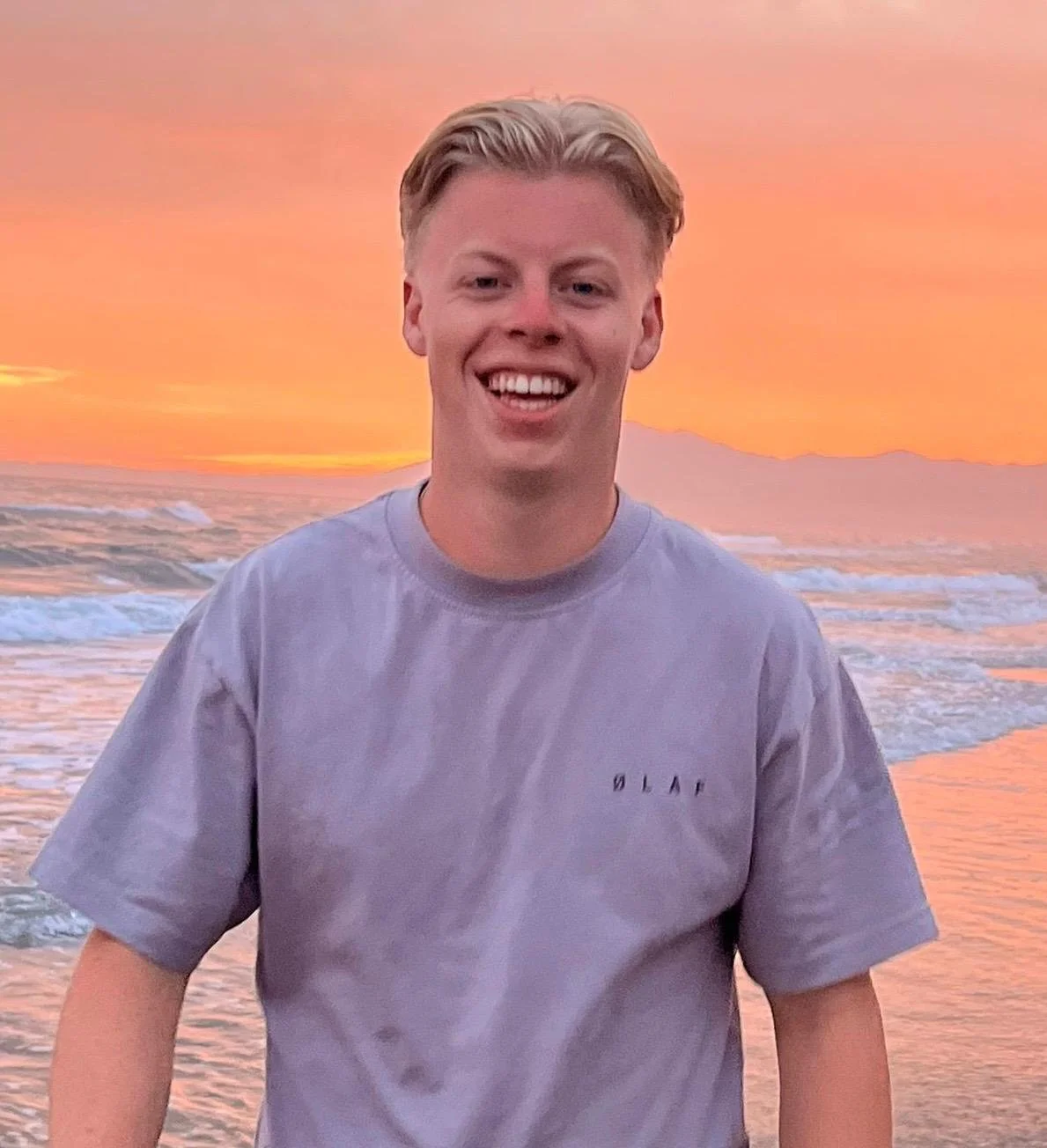 Een jonge man staat op het strand tijdens zonsondergang, lachend en kijkend naar de camera.