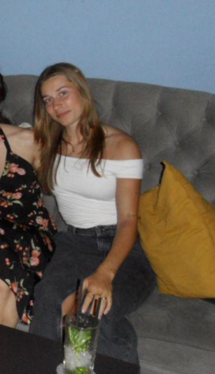 Een jonge vrouw met lang bruin haar die op een bank zit, met een gele kussen op haar rechterkant.