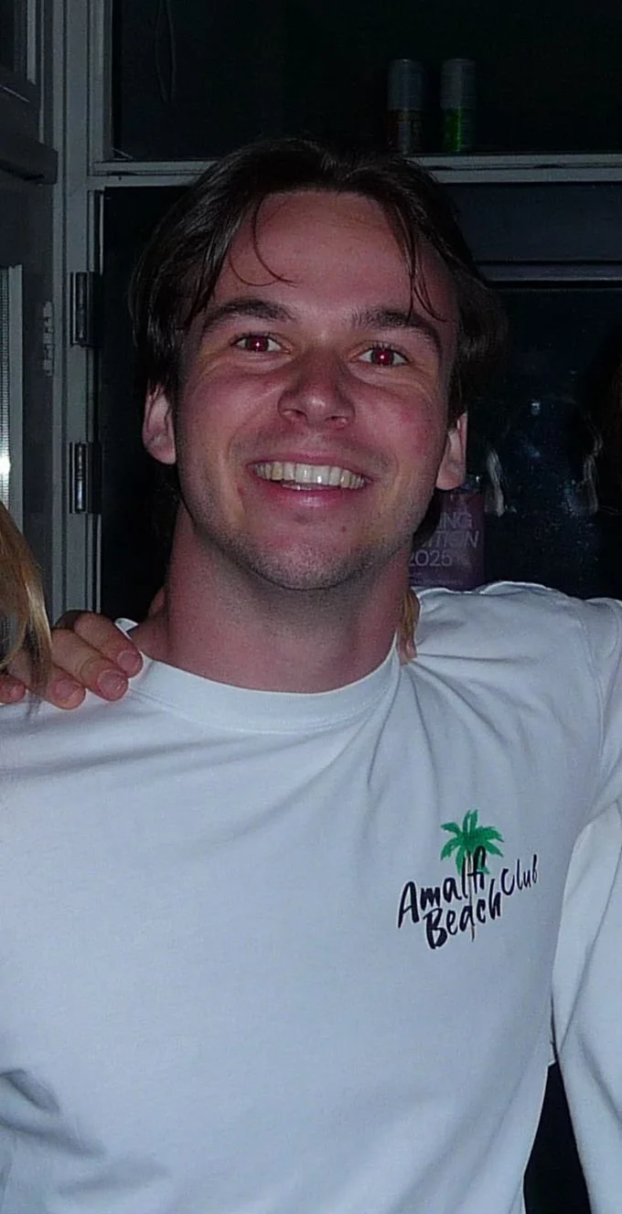 Een lachende man met donkerbruin haar draagt een witte T-shirt met de tekst 'Amalfi Beach Club' en een groene palmboom. De achtergrond lijkt een kraakheldere, zonnige locatie.