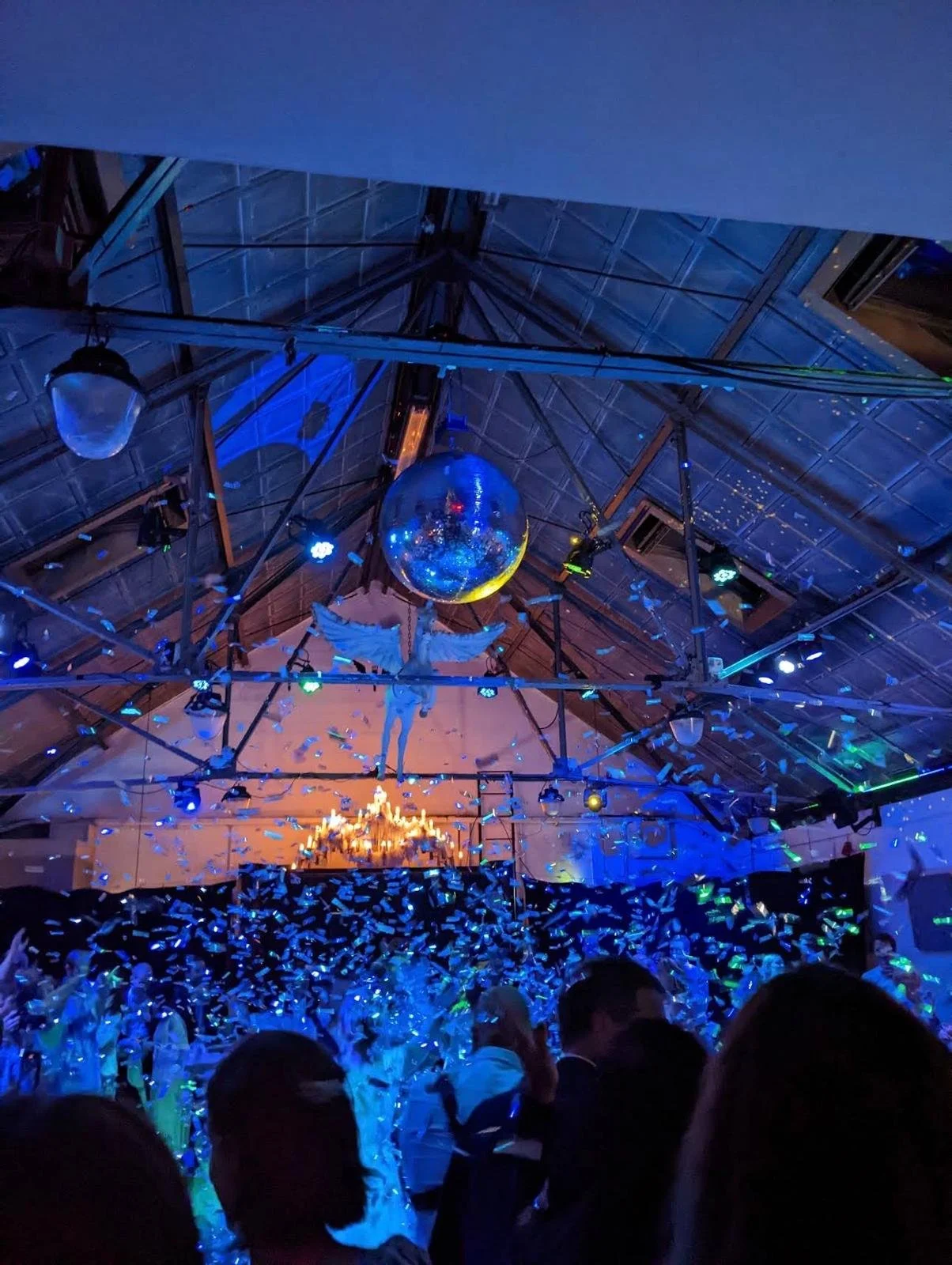 Binnenzaal met confetti, blauwe en gekleurde lichten, een grote spiegelbol, en een dansvloer vol mensen tijdens een feest of evenement.