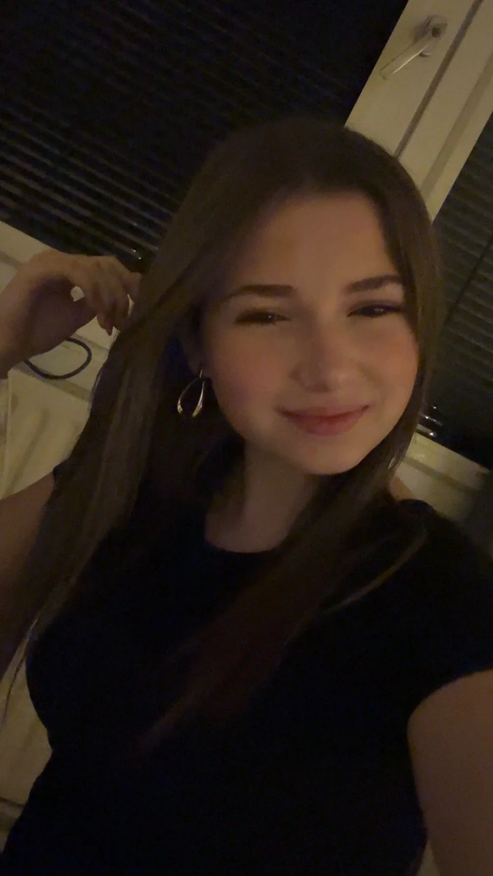 Een vrouw met bruin haar, die glimlacht en oorringen draagt, neemt een selfie in een dim verlichte kamer.