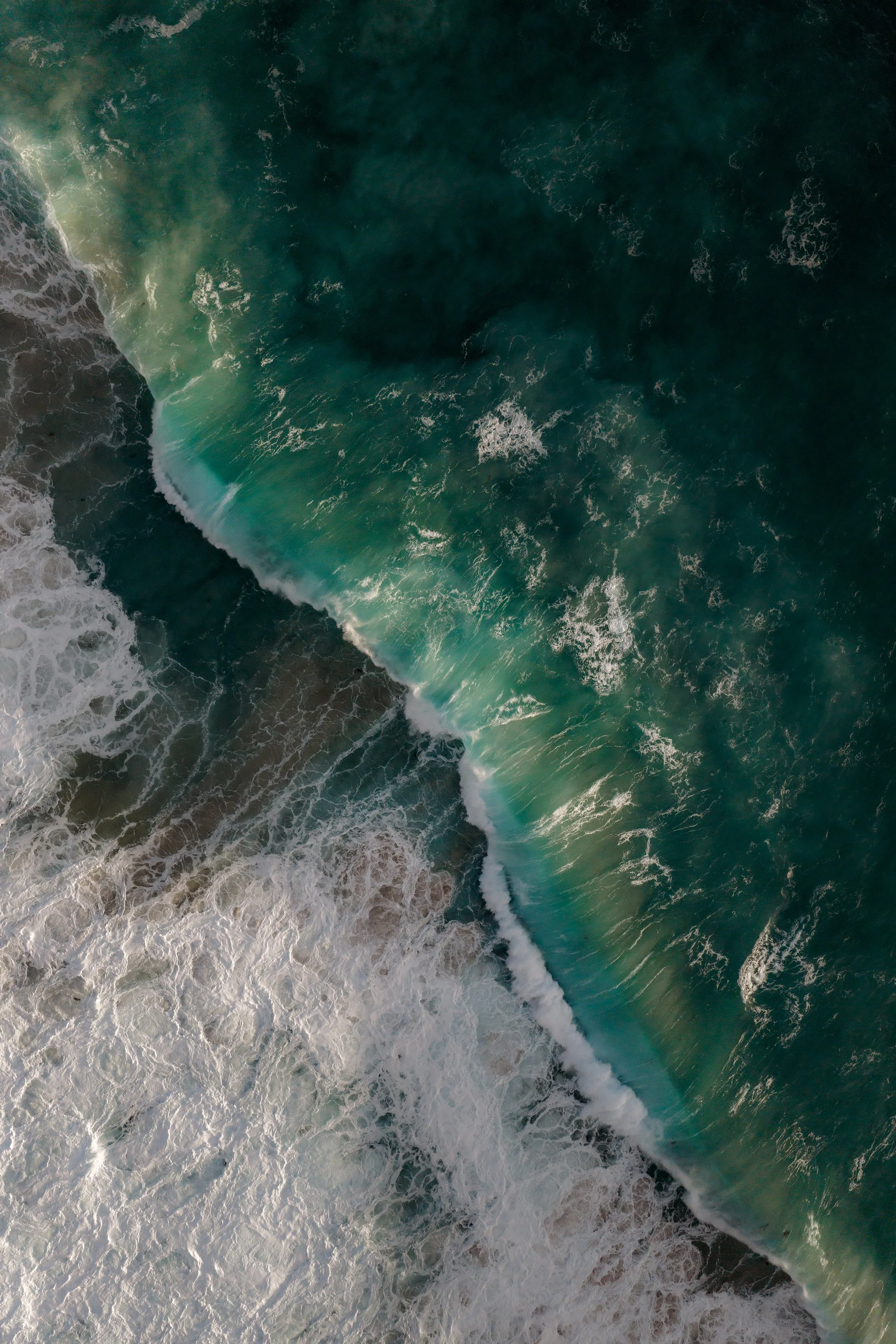 An aerial view of the ocean showing waves crashing onto the shore with foam and a large rock formation.