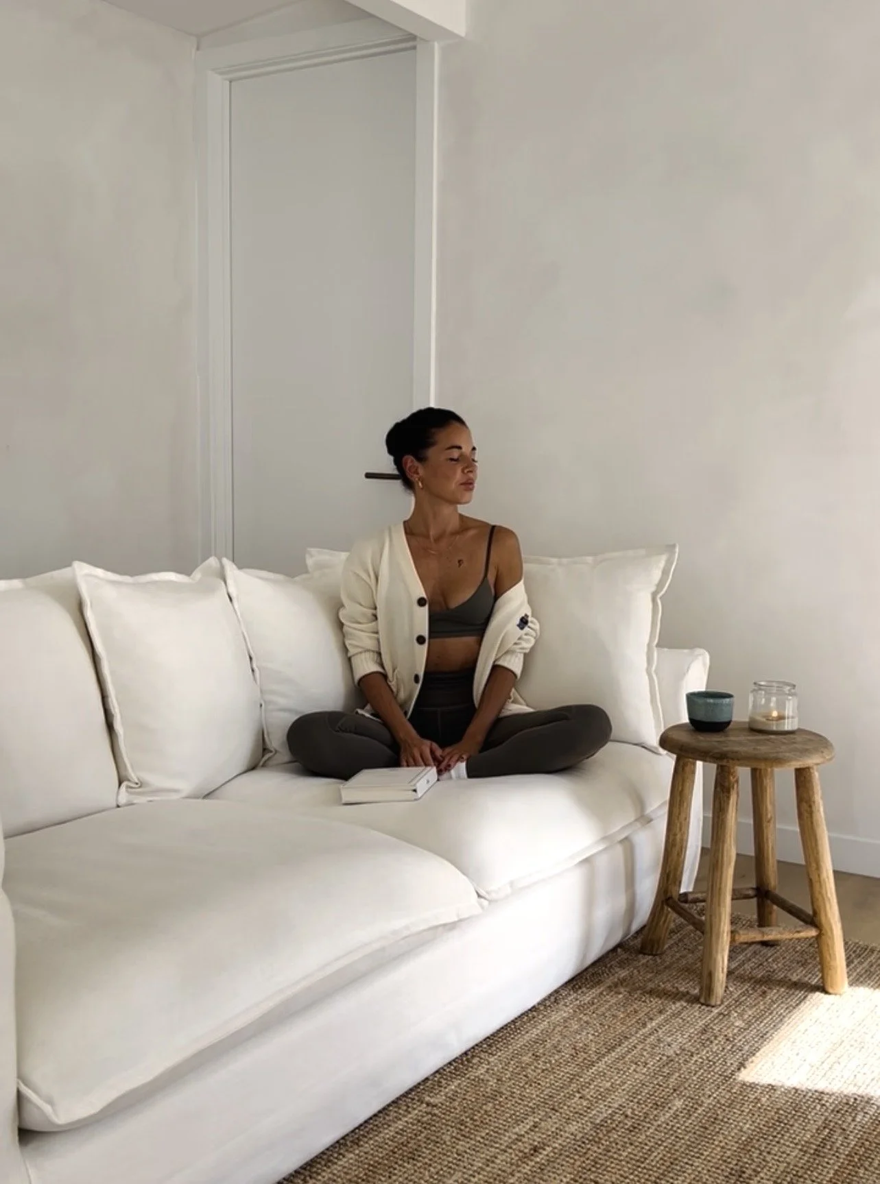 A woman sitting cross-legged on a white couch in a minimalist living room, with a closed book on her lap, next to a small wooden side table holding a candle, a bowl, and a glass jar, against a plain white wall. Peace, gratitude, slow morning rituals.