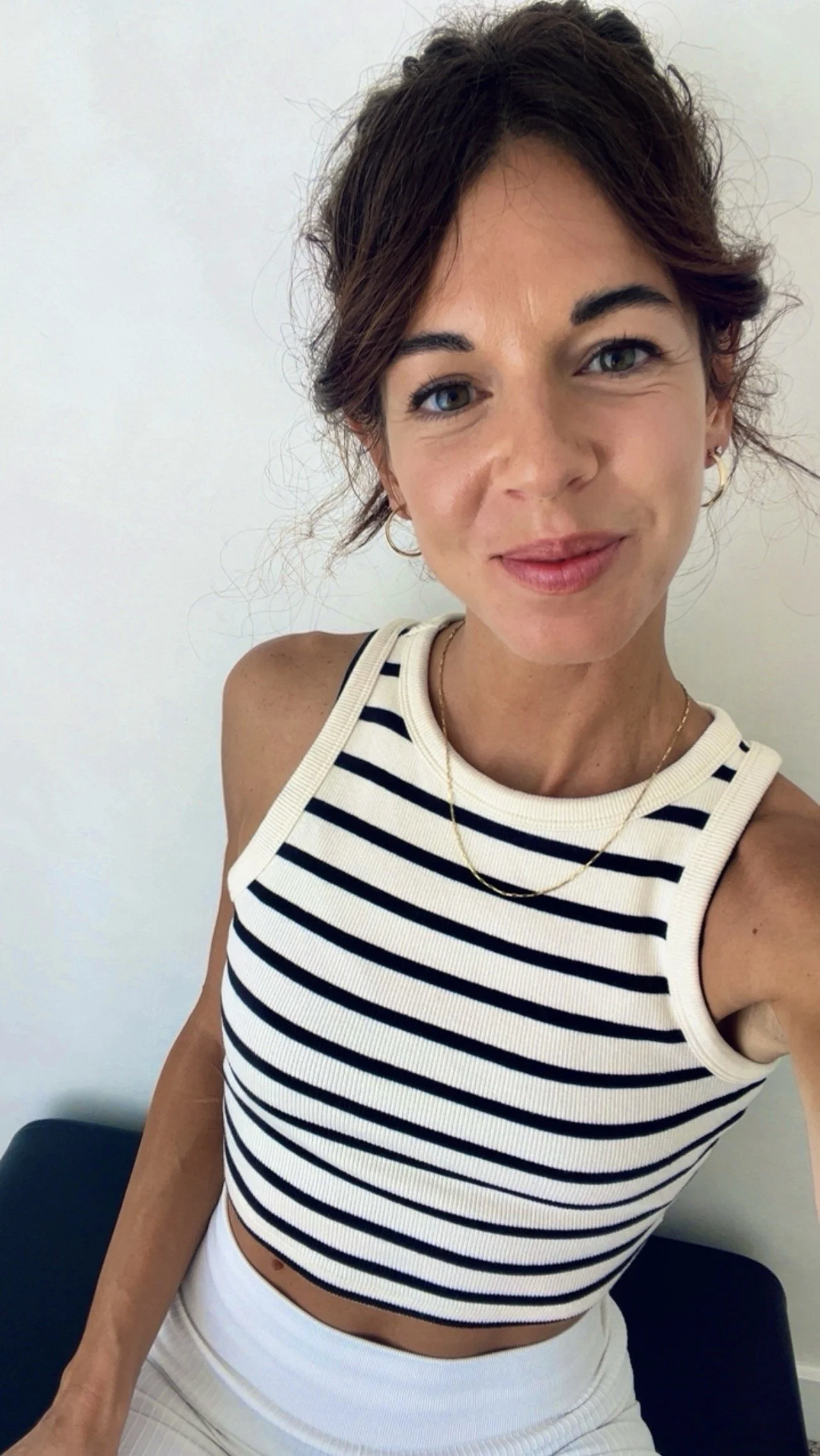 A young woman with short, wavy brown hair and blue eyes, wearing a sleeveless striped top and white pants, posing for a selfie against a plain white wall. Founder of ILIO Club, happy, serene, calm because she is becoming that elevated version of hers