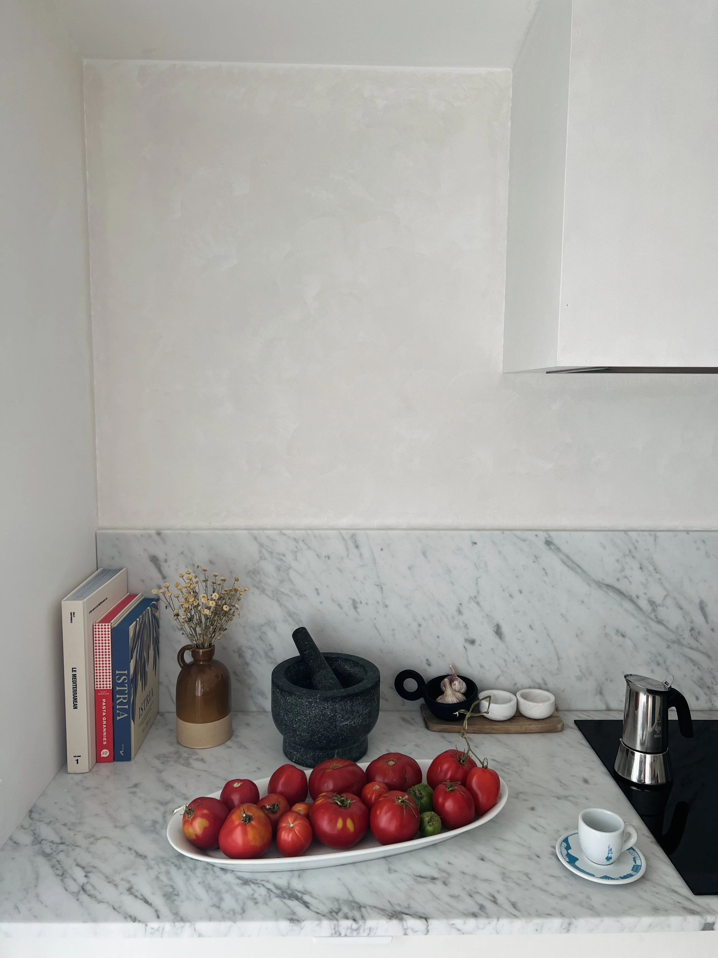 A kitchen counter with a plate of homegrown tomatoes, a bowl of garlic, a mortar and pestle, a plant in a vase, several cooking books, a small espresso cup on a saucer and an italian stovetop coffee machine.  Homecooking staples, slow living.