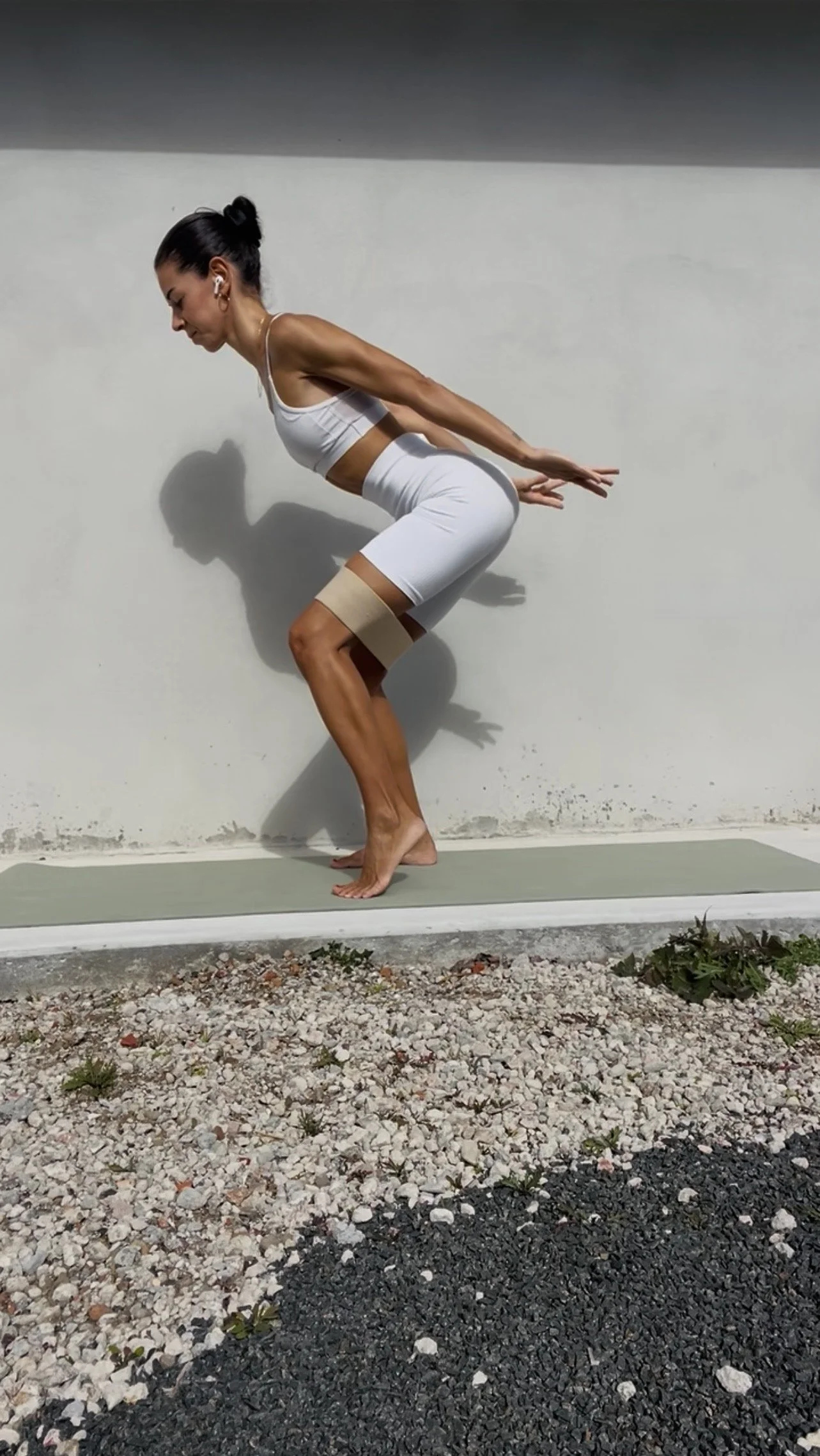 A woman practicing pilates outdoors on a green mat against a white wall, wearing a white sports bra and shorts, with her hair tied back and headphones in her ears. Pilates practice, anytime, anywhere.