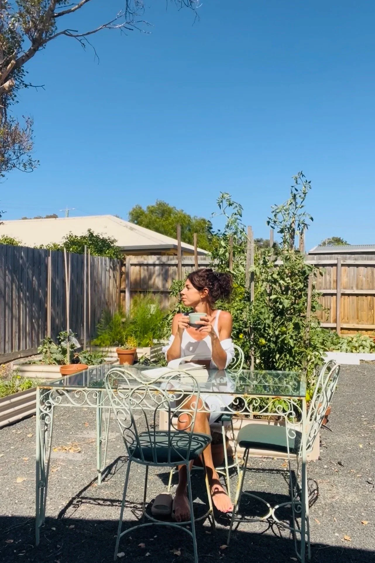 A woman sitting outdoors at a garden table, holding a cup, in her vegetable garden, under clear blue sky. Taking the time to reflect, soak the sun, journal, sipping matcha, listening to nature.