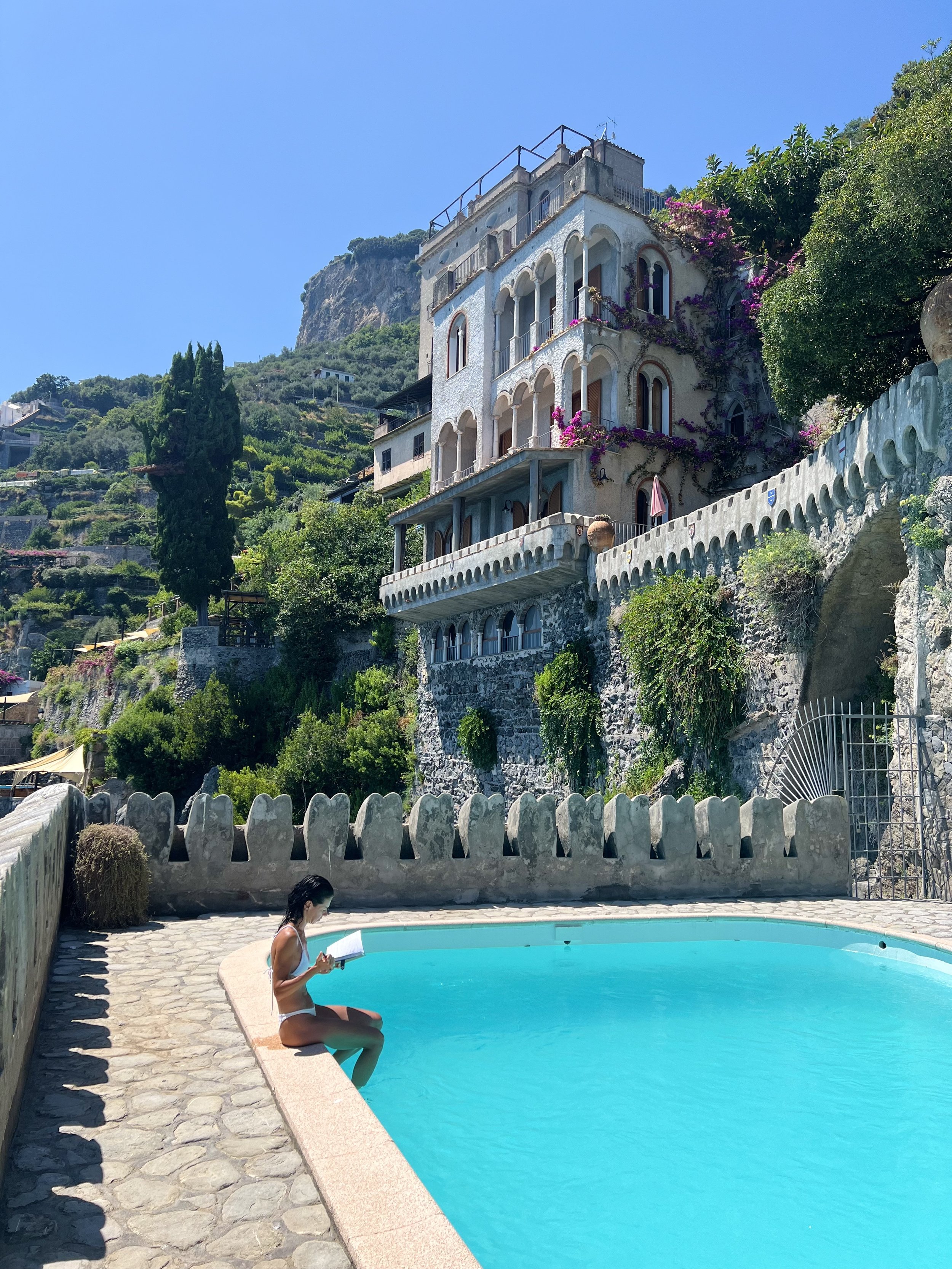 A woman in a white bikini sitting on the poolside reading a book, with a house and hillside in the background. Amalfi coast, one of the most perfect corners of Italy. Slow living in travelling, travelling with intention.