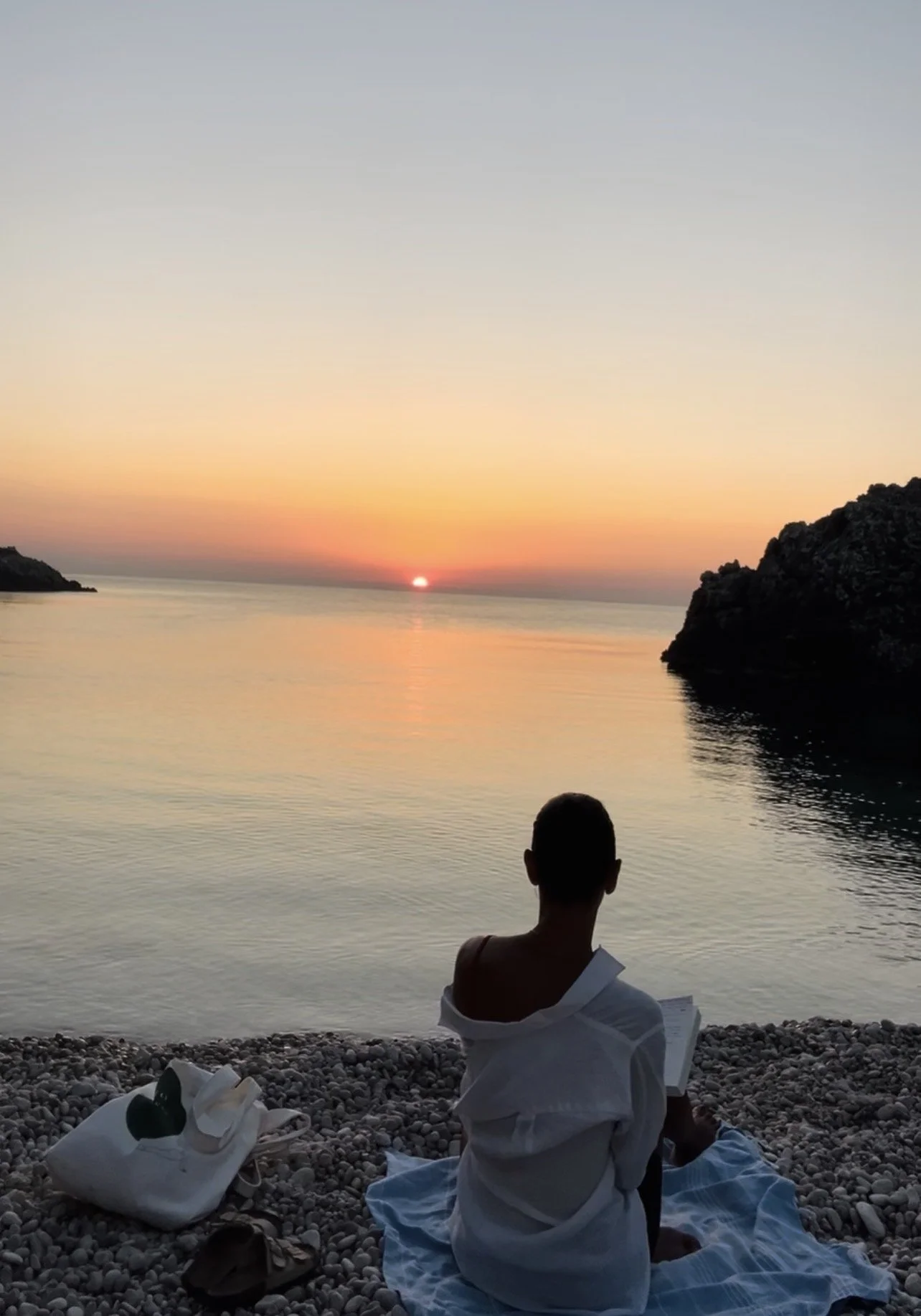 Person sitting on a pebble beach, facing the ocean, watching a sunrise with colorful sky and calm water, surrounded by rocky cliffs. Taking the sunrise, the sea, the silence in, before the noise of the day start.